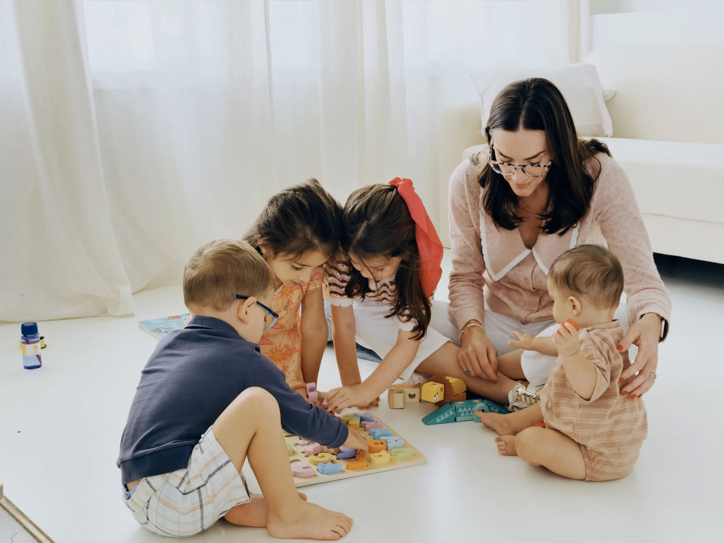A woman and several children playing a colorful board game on the floor in a bright room with white curtains.