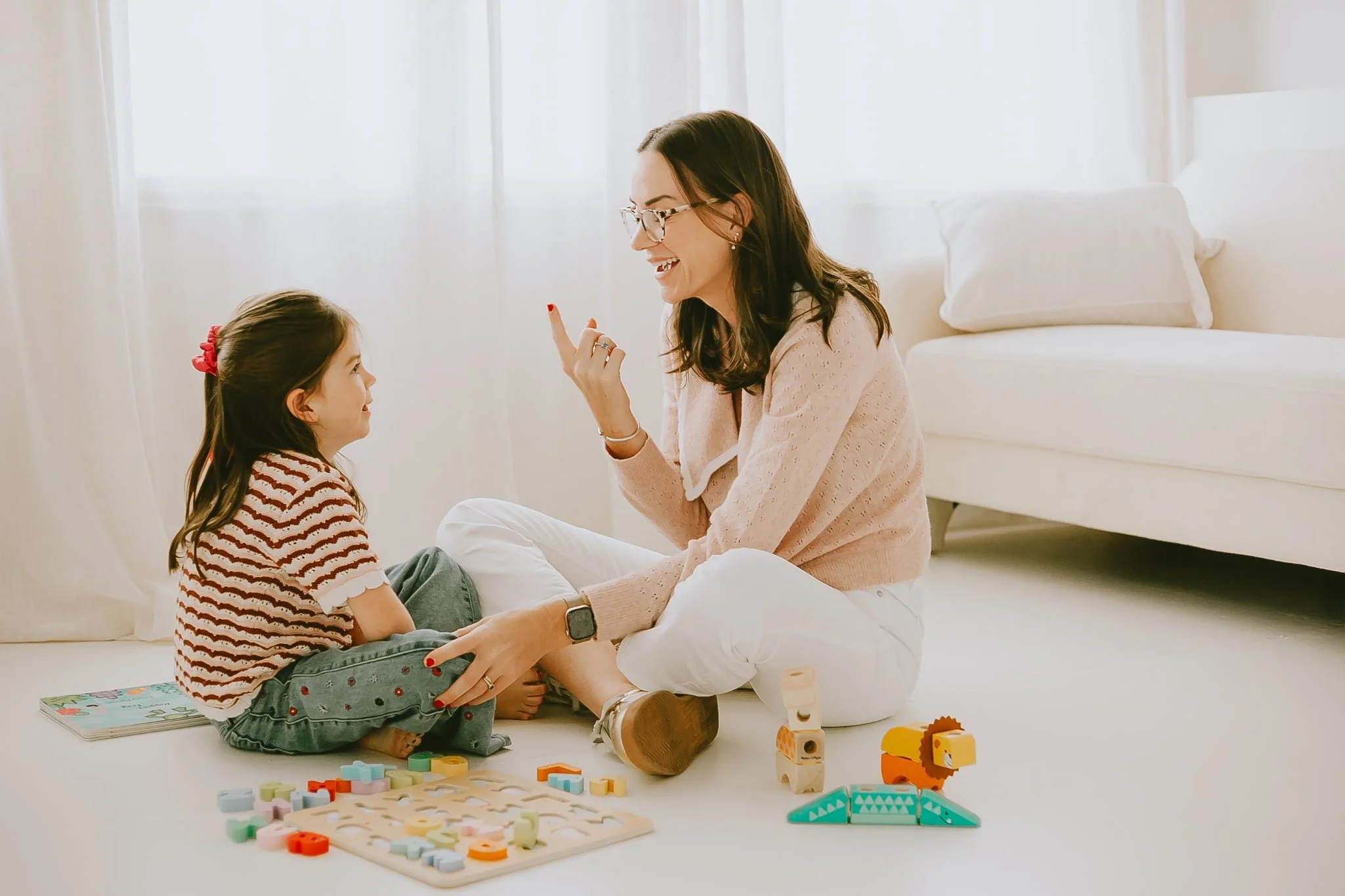 A woman and girl playing with colorful wooden toys and puzzles on the floor of a bright room. The woman is smiling and gesturing with her finger as she interacts with the girl, who looks up at her. There are toys and a book scattered around them.