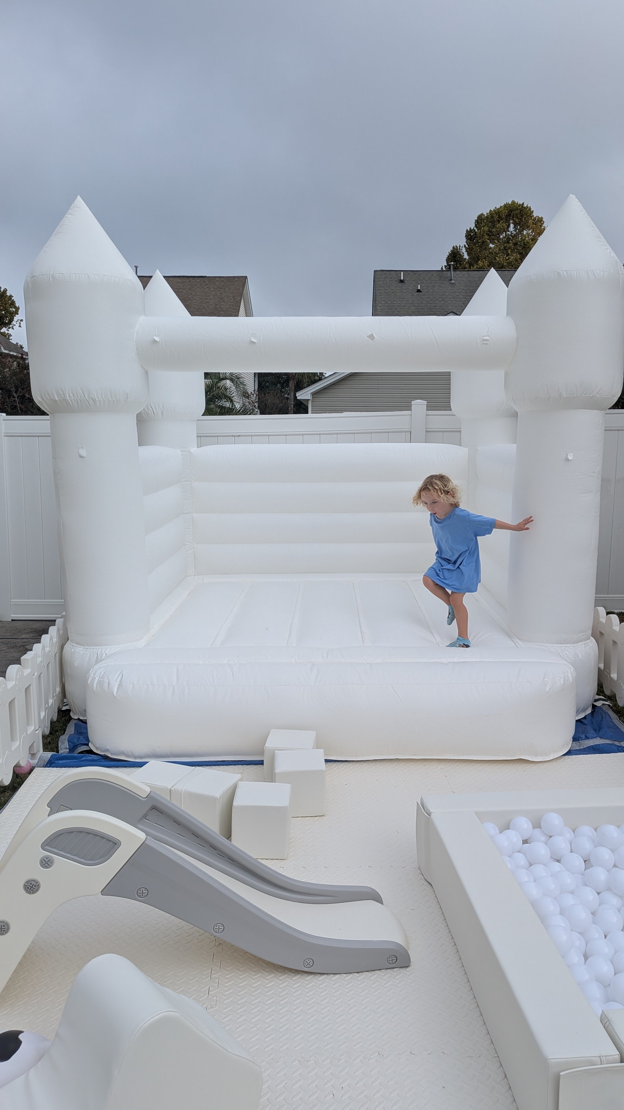 A young girl in a blue dress playing on a large white inflatable castle with a slide and a ball pit outside in a backyard with a white fence and houses in the background.