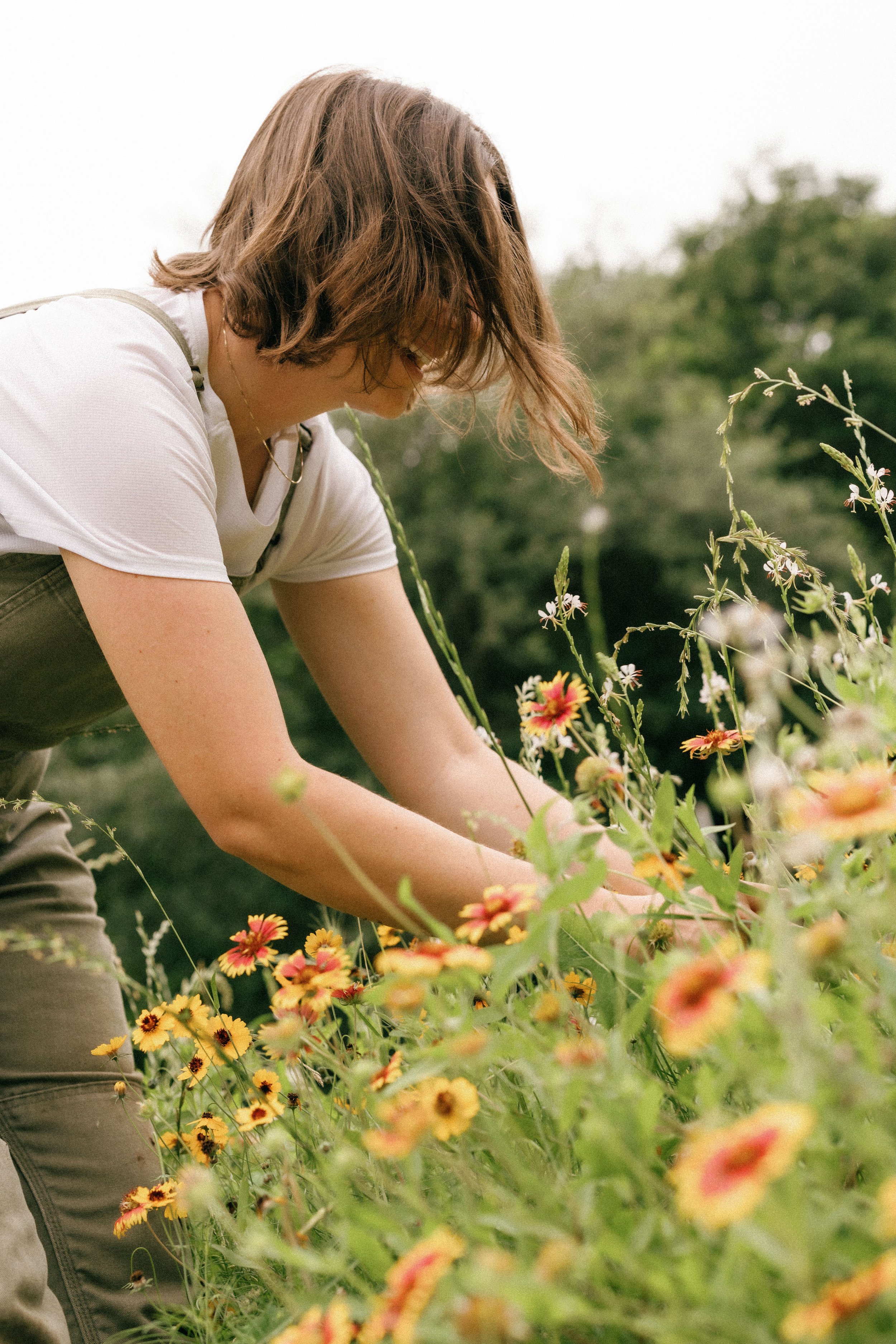 A woman with light brown hair, wearing a white t-shirt and khaki pants, is tending to a garden filled with colorful flowers. She is bending over and appears to be planting or tending to the flowers, with a blurred green background of trees and foliage.
