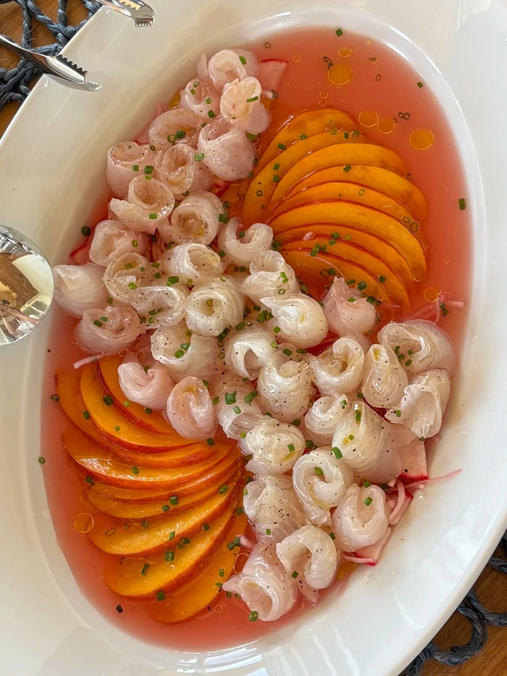 A white bowl containing a seafood dish with diced pink seafood, sliced yellow and orange bell peppers, and green chopped herbs in a pink soup or sauce.