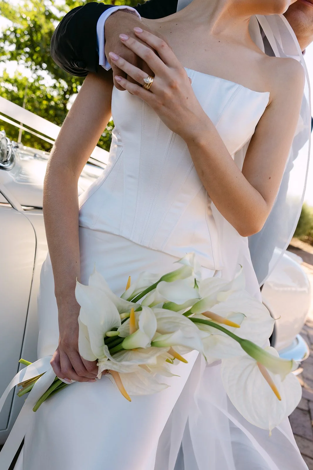 A bride in a white gown holding a bouquet of white calla lilies, with her left hand resting on her chest, showing a wedding ring. There is a vintage car and green foliage in the background.