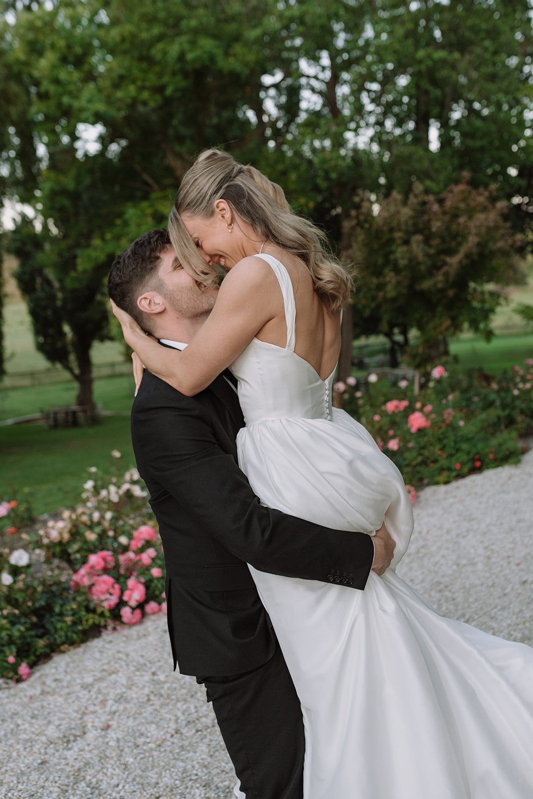 A man in a black tuxedo lifts a woman in a white wedding dress, hugging and smiling affectionately in a garden with blooming pink and white roses and green trees.