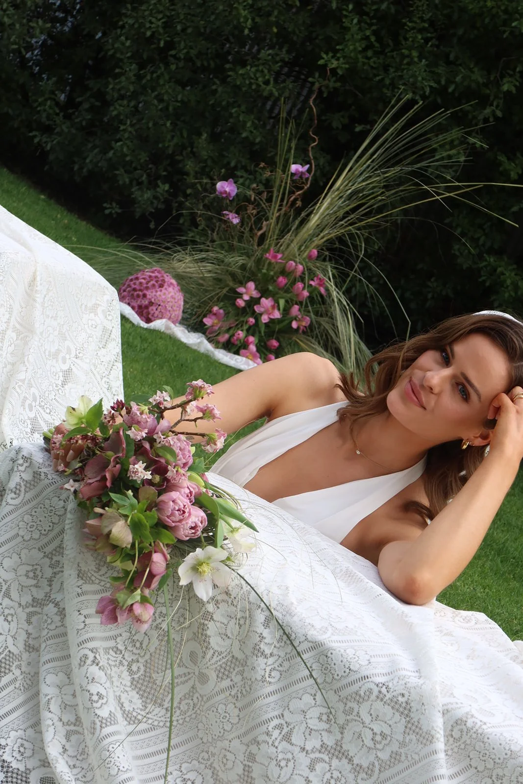 A woman in a white dress lounging outdoors at a table with lace cloth, surrounded by pink and white flowers, with greenery in the background.