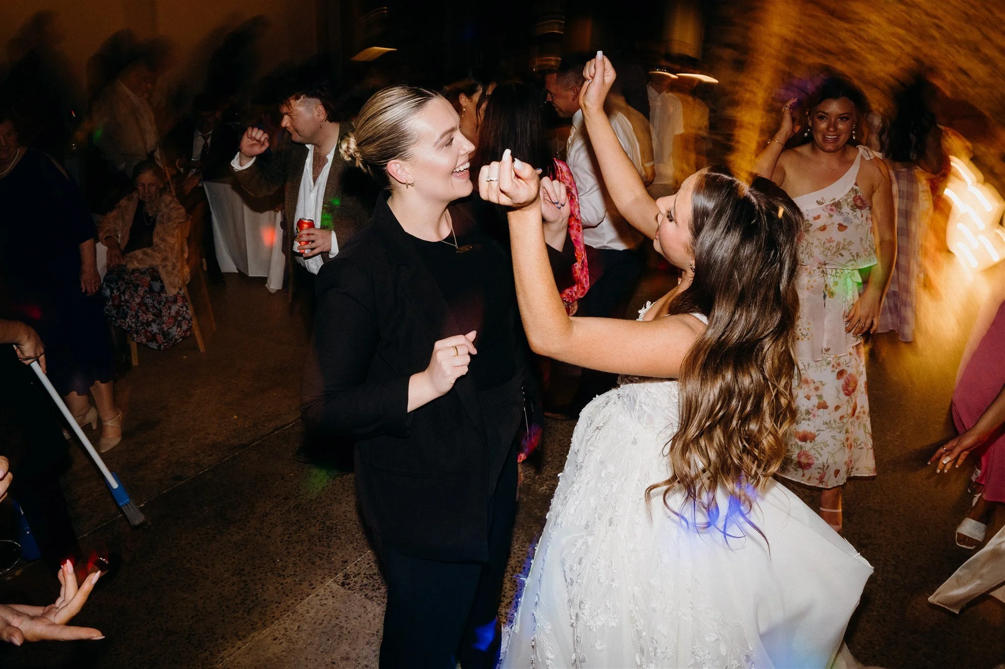 People dancing and celebrating at an indoor wedding reception, including a woman in a white wedding dress and a woman in a black dress smiling and enjoying music.