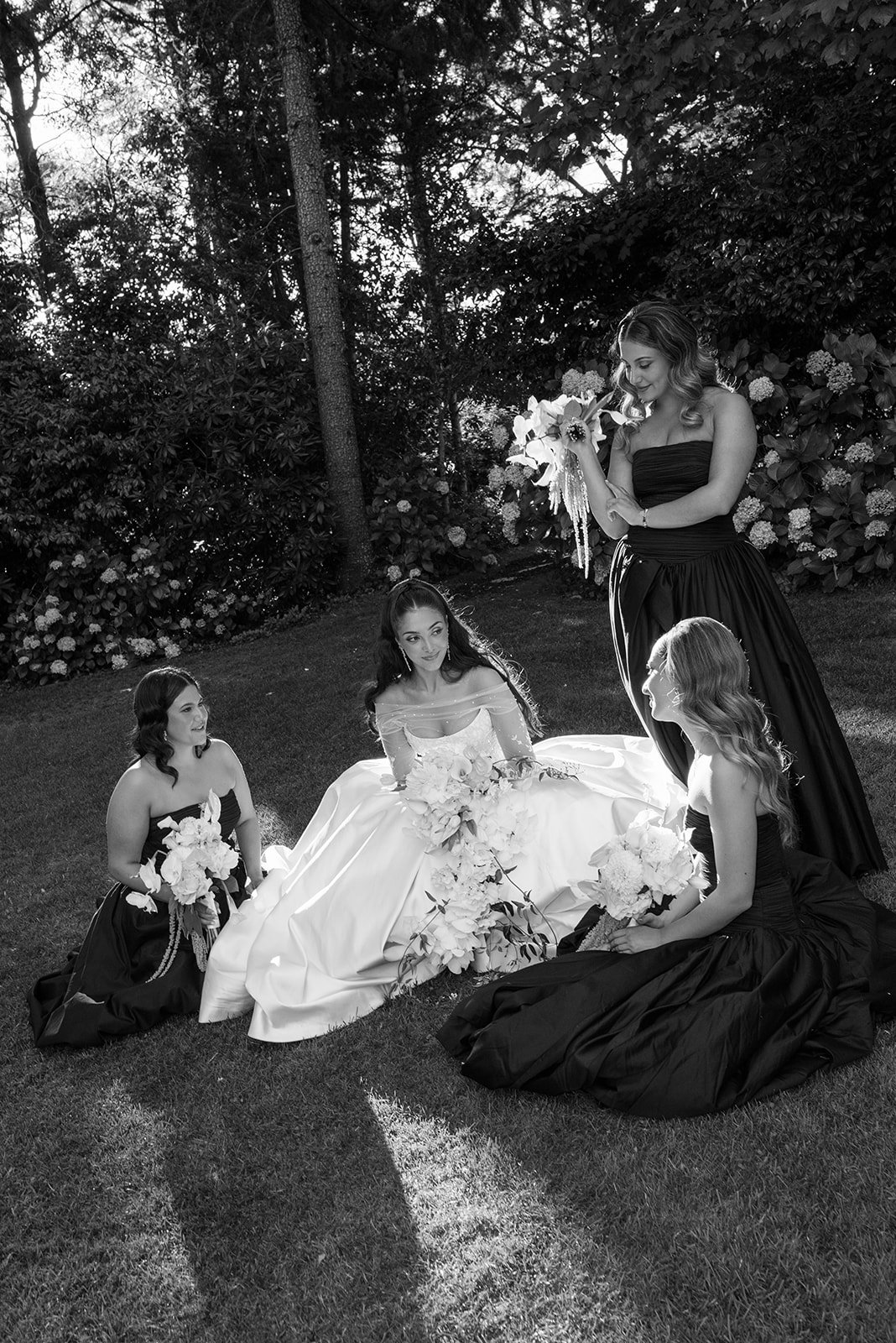 A black and white photo of a woman in a wedding dress seated on the grass, surrounded by three women in dark dresses, all holding bouquets. One woman standing is holding a bouquet and looking at it. The background has trees and bushes.