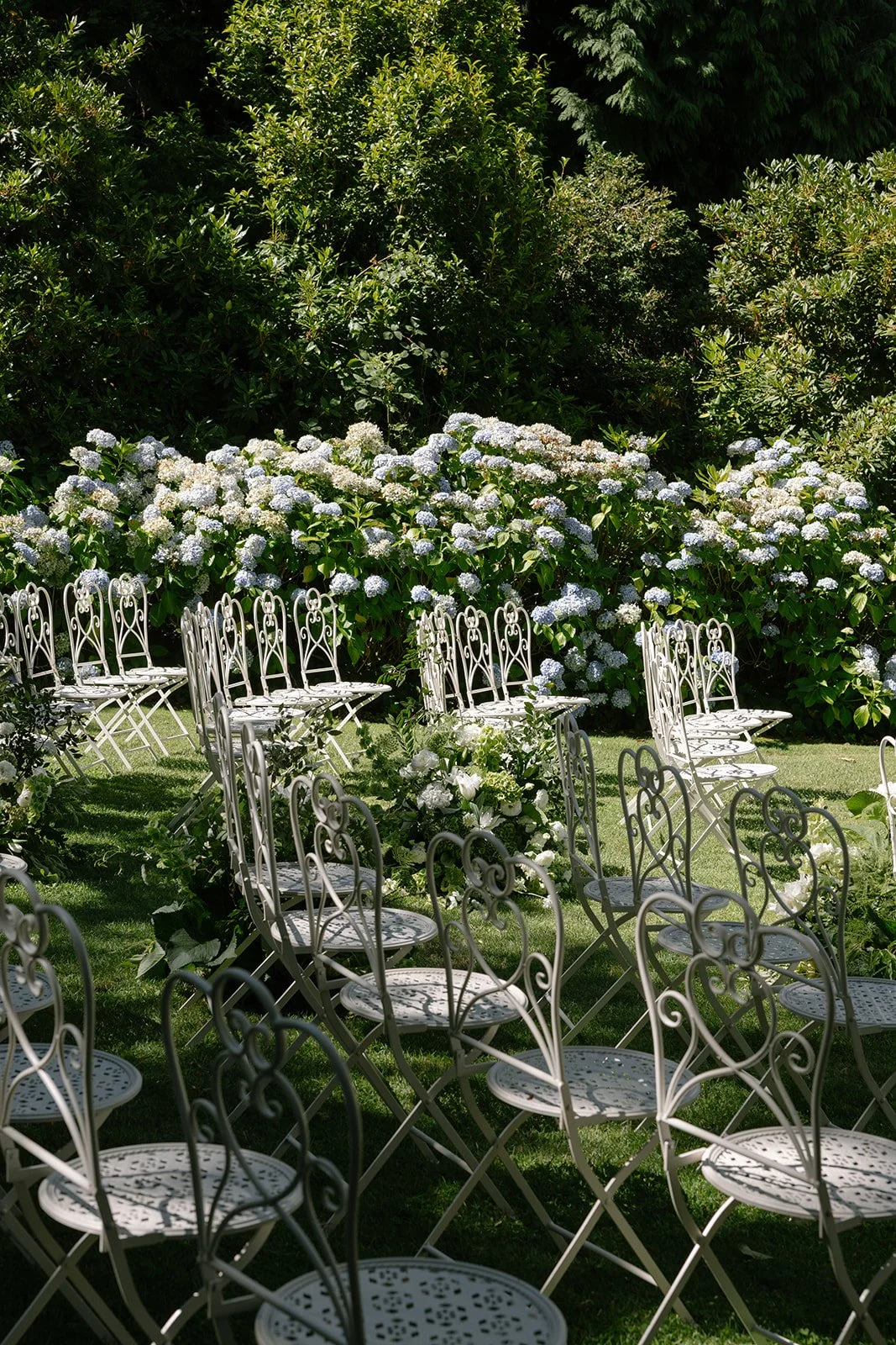 White metal chairs arranged outdoors in a garden setting with lush green bushes and white flowering plants in the background.