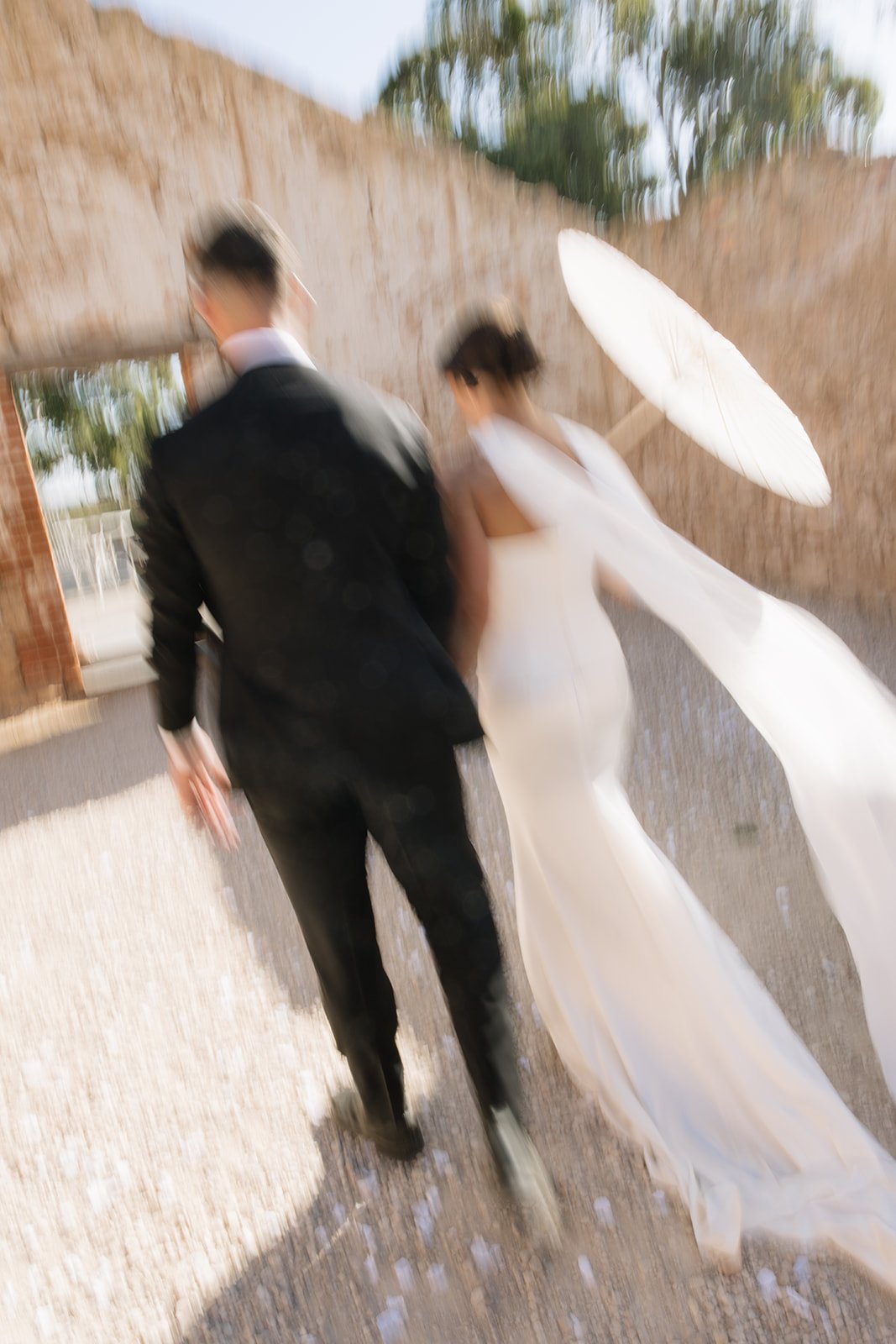 A couple in wedding attire walking outdoors, with the bride holding a large white umbrella.