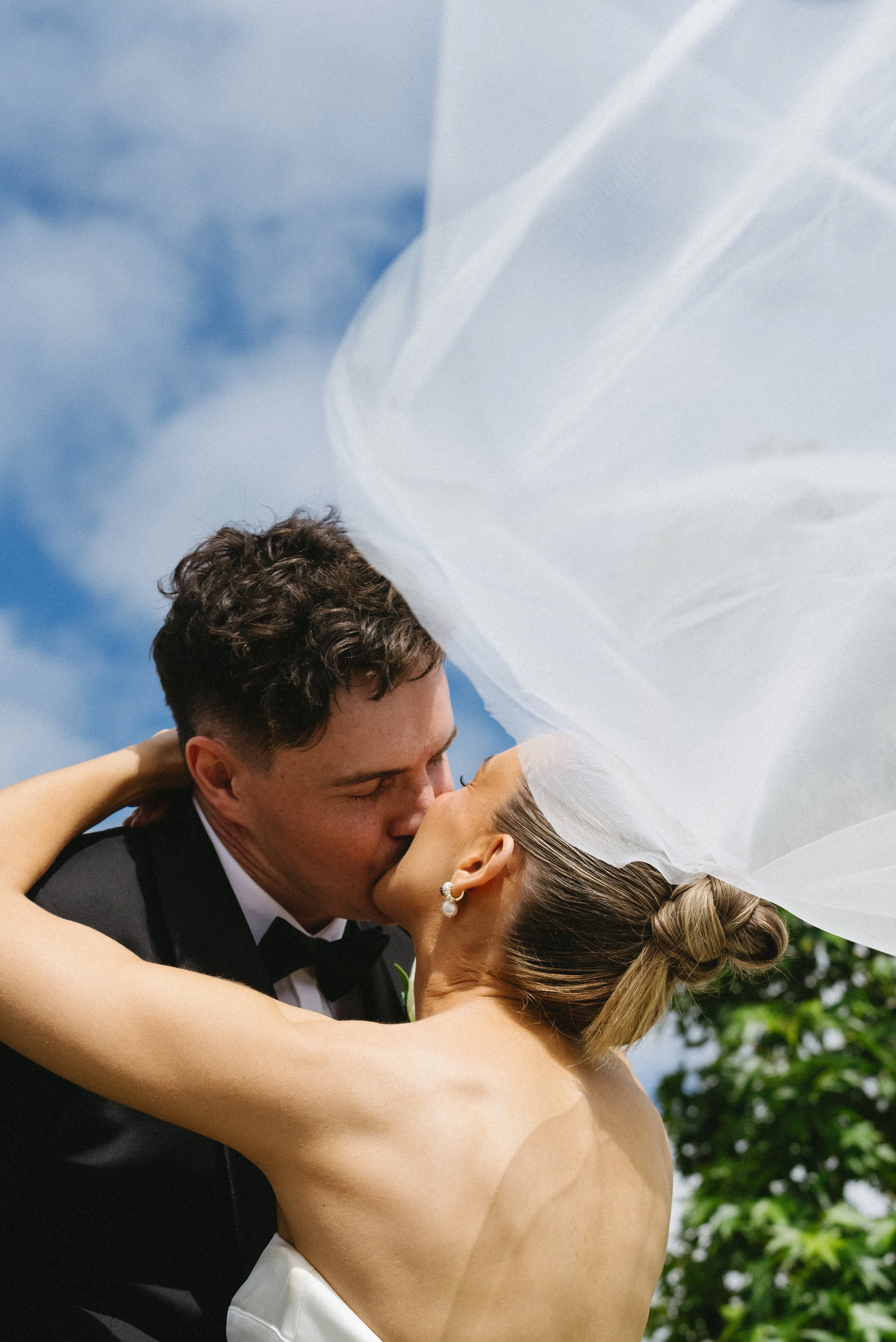 A couple sharing a kiss at their wedding, outdoors with a bright sky and green foliage in the background, the bride wearing a white dress and earrings, the groom in a black tuxedo.