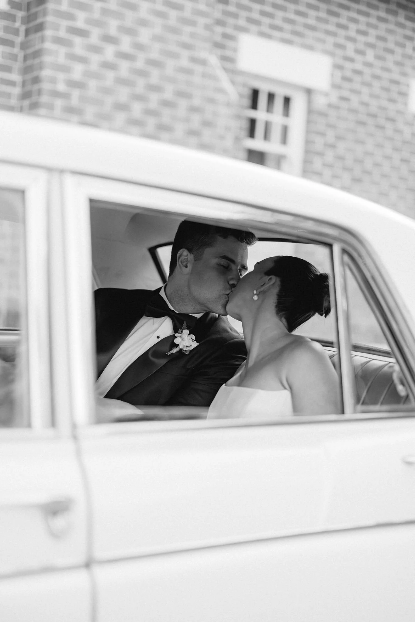 A black and white photo of a man and woman sharing a kiss inside a vintage car, with a brick building visible in the background.