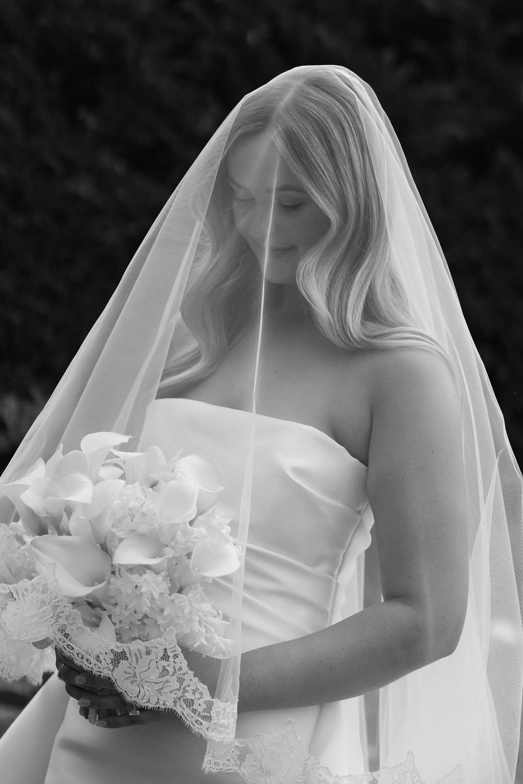 A bride in a strapless wedding dress with long wavy hair, holding a bouquet of flowers, with a veil over her head, smiling softly in an outdoor setting.