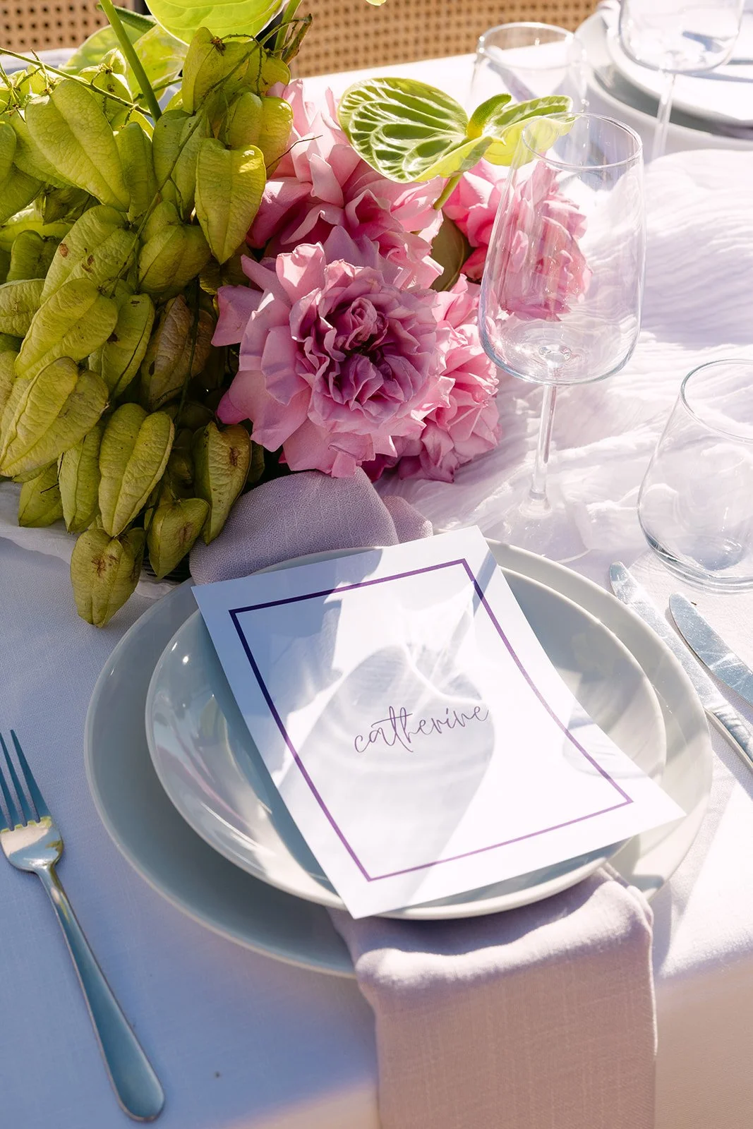 A table setting with a white plate, a menu with the name Catherine, a bunch of pink flowers, green leaves, a water glass, an empty wine glass, a fork, and a knife. The table has a white tablecloth and a pink napkin.