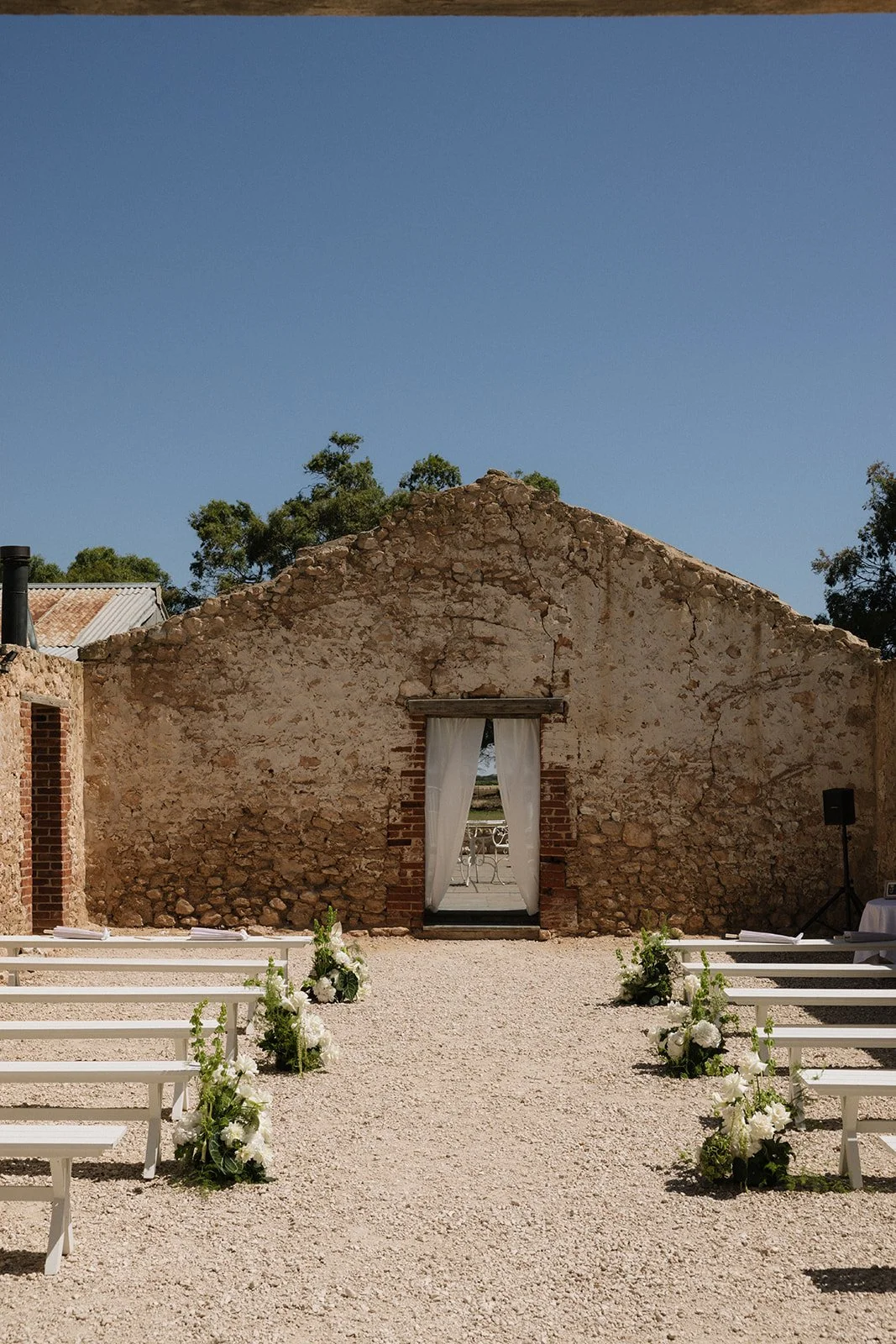 An outdoor wedding setup with white benches and floral arrangements on a gravel path leading to an old stone building with a door and white curtains.