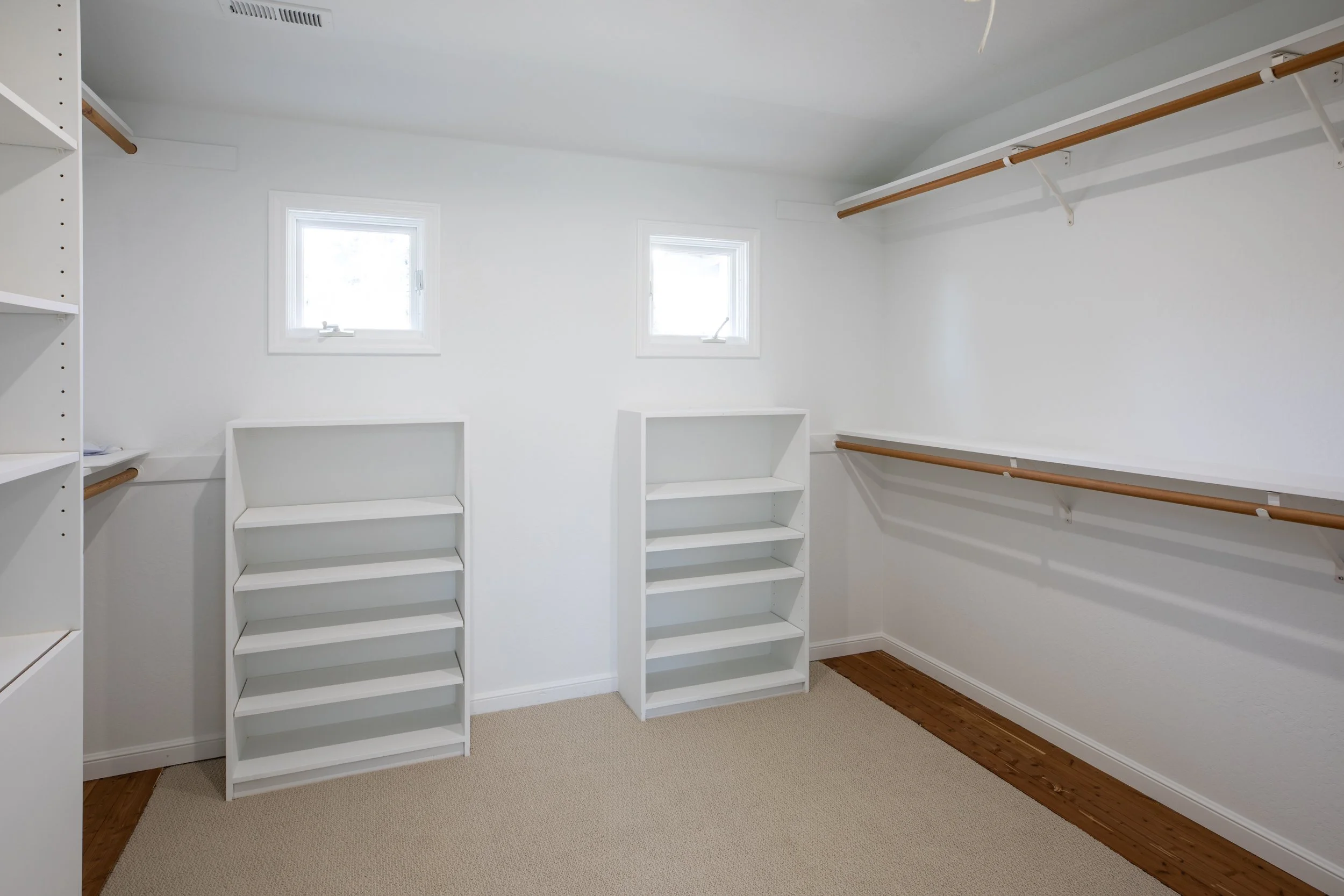 Empty walk-in closet with white shelves and hanging rods, two small windows, beige carpet, and wooden flooring along the wall.