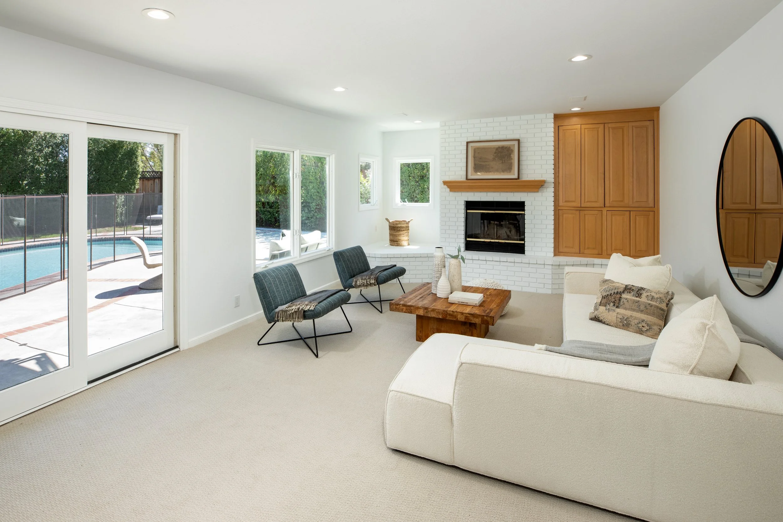 Living room with white walls, a white brick fireplace with a wooden mantle, and multiple windows letting in natural light. Outside, a pool and patio area are visible through the glass door.