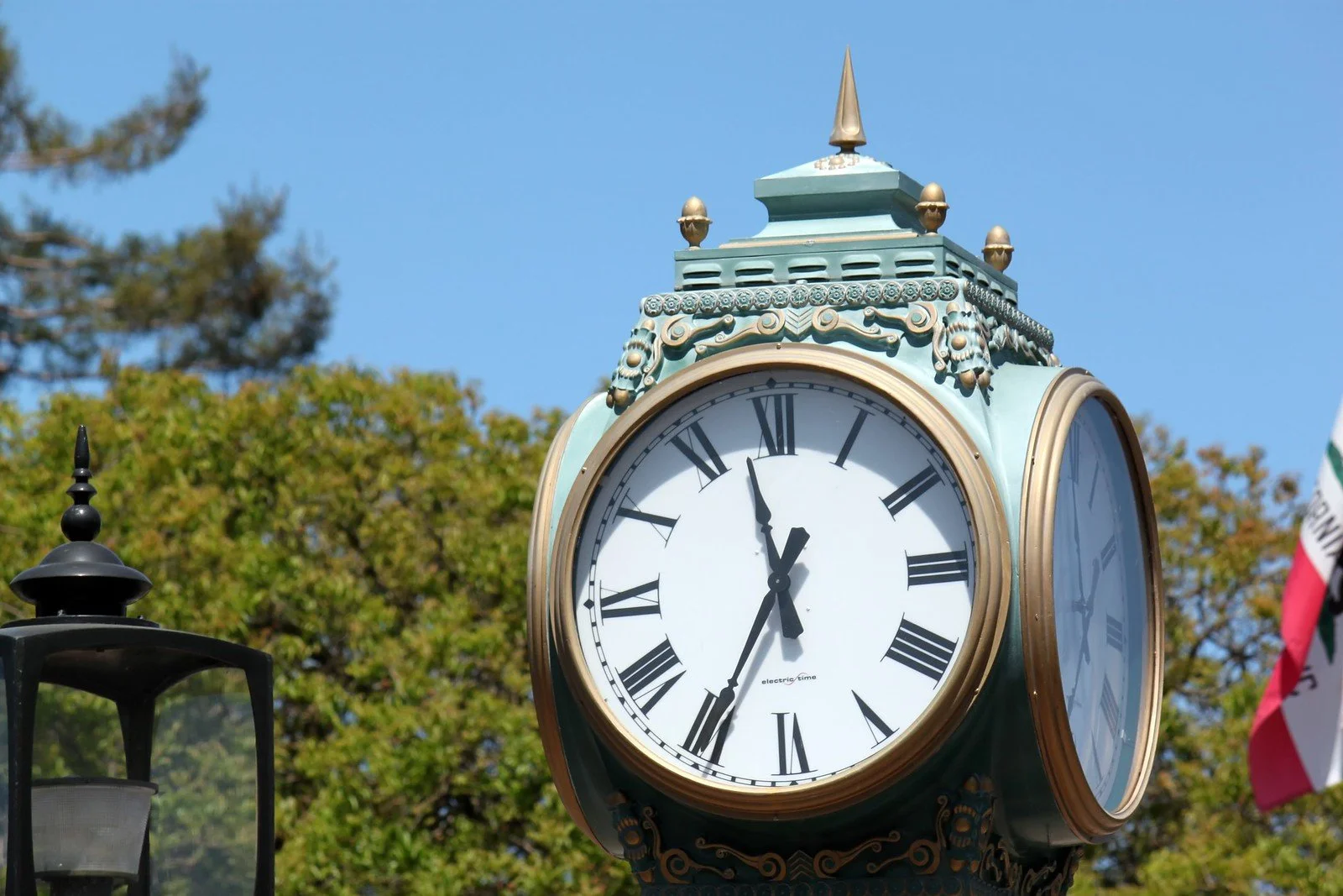 An ornate clock showing the time as approximately 12:02, mounted outdoors with trees and a clear blue sky in the background.