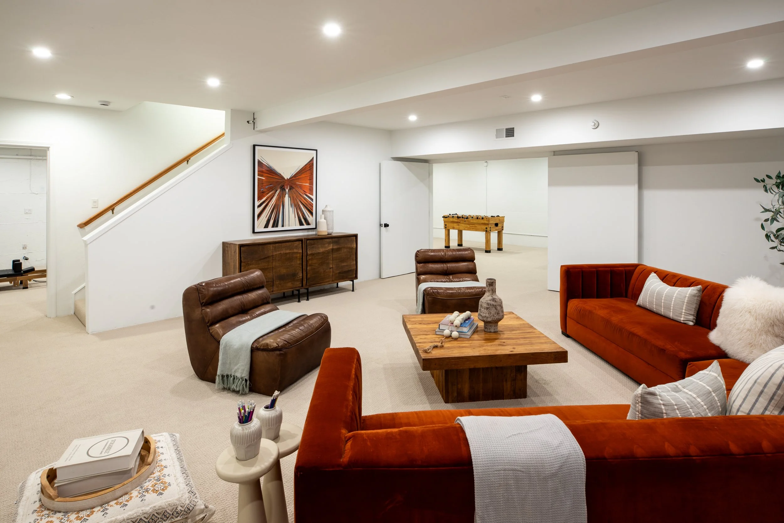 Living room with white walls, beige carpet, red velvet sofas, brown leather chairs, a wooden coffee table, decorative vases, artwork, a foosball table in the background, and recessed ceiling lights.