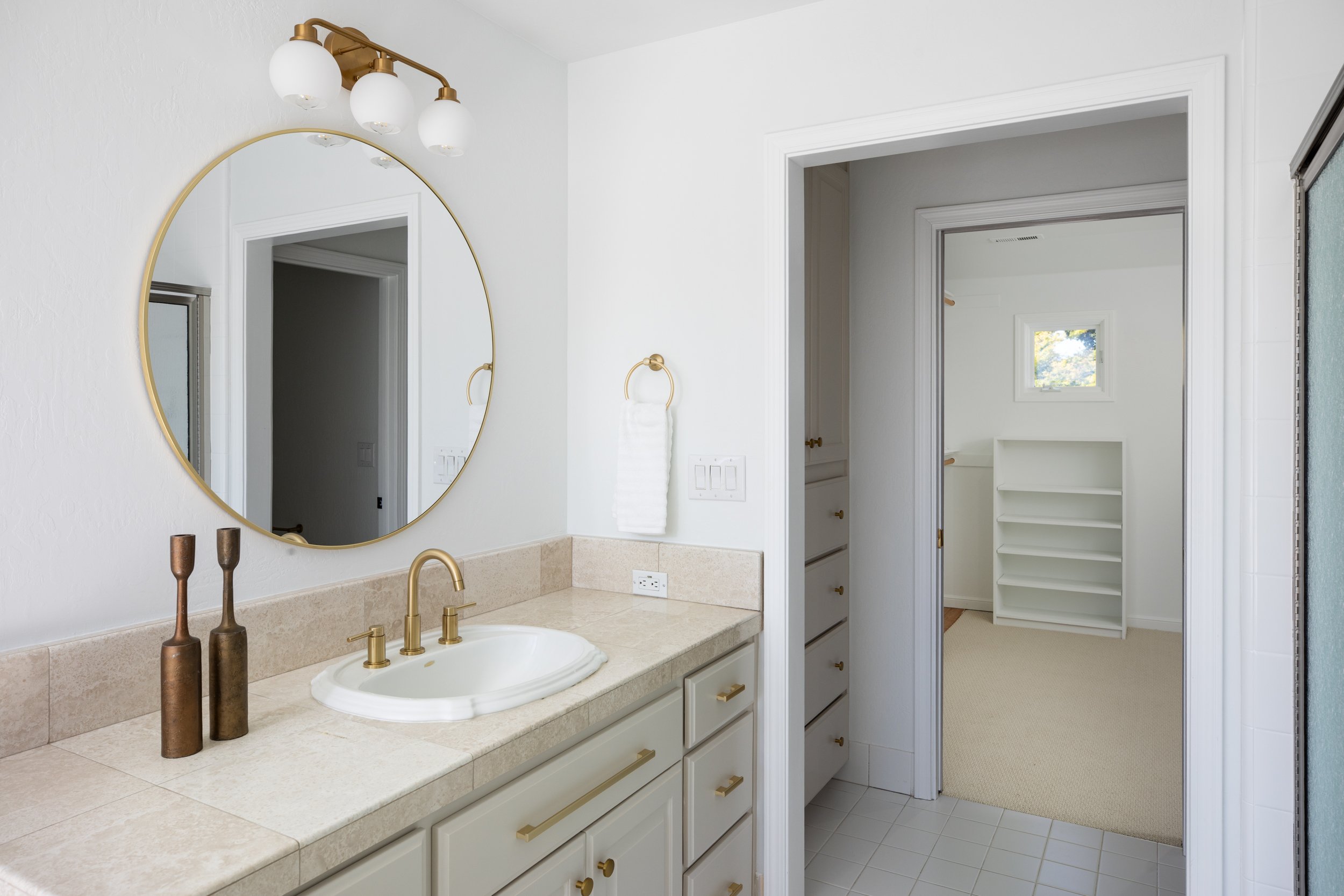 A bathroom with a beige countertop, a white sink, and a large round mirror with a gold frame. There are three decorative vases on the countertop, a gold faucet, and a wall-mounted light fixture with four white globe lights. The walls are white, and t