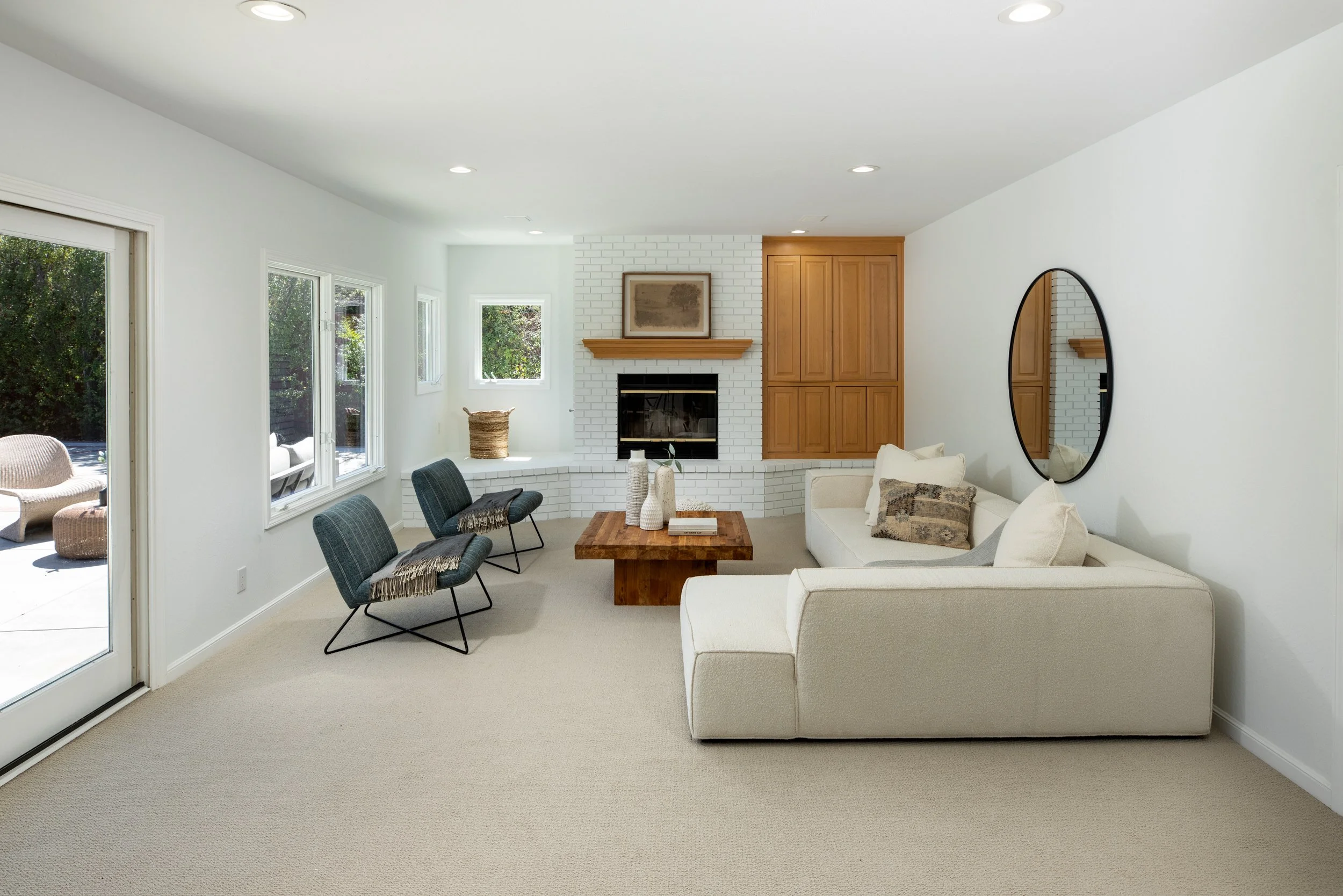 Living room with white walls, beige carpet, white sofa, blue chairs, wooden coffee table, fireplace with white brick surround, flat-screen TV, built-in wooden cabinets, window seats with wicker baskets, wall mirror, and large sliding glass door leadi