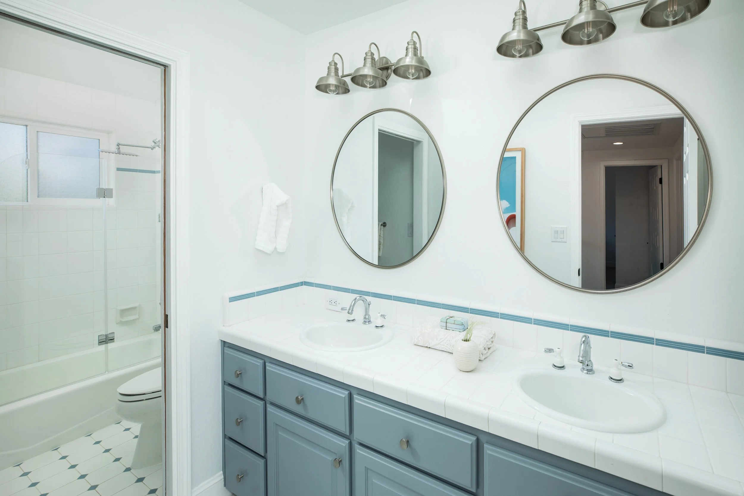 A bathroom with a blue vanity, white tiled countertop, two oval mirrors, and a light fixture with six metal lamps. There is a shower with frosted glass doors and a small window in the shower area.