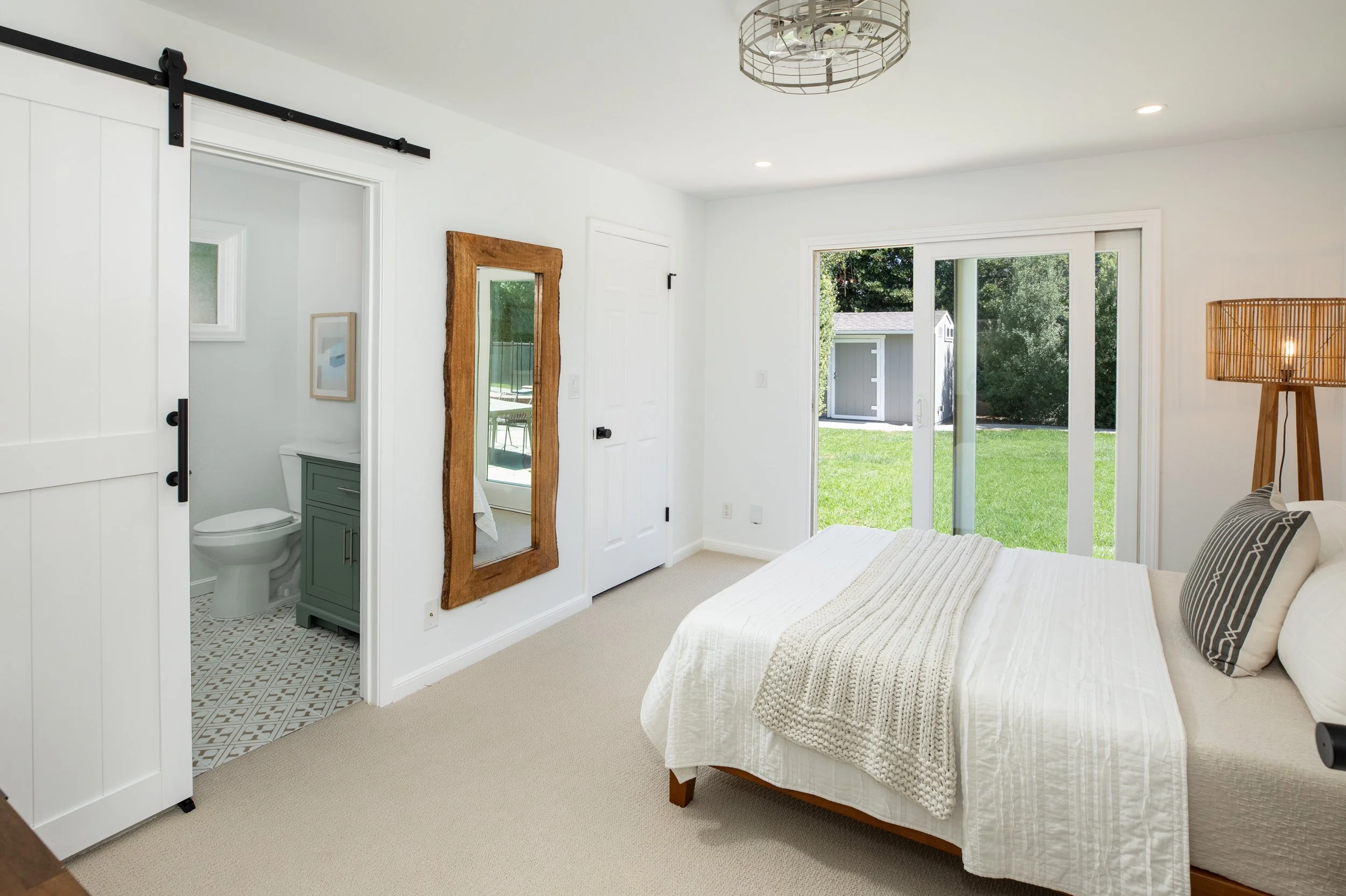 A bright bedroom with a bed covered in a white quilt, a striped pillow, and a wooden lamp. Large sliding glass doors lead to a green backyard. A full-length mirror with a wooden frame leans against the wall near a small bathroom with a toilet and gra