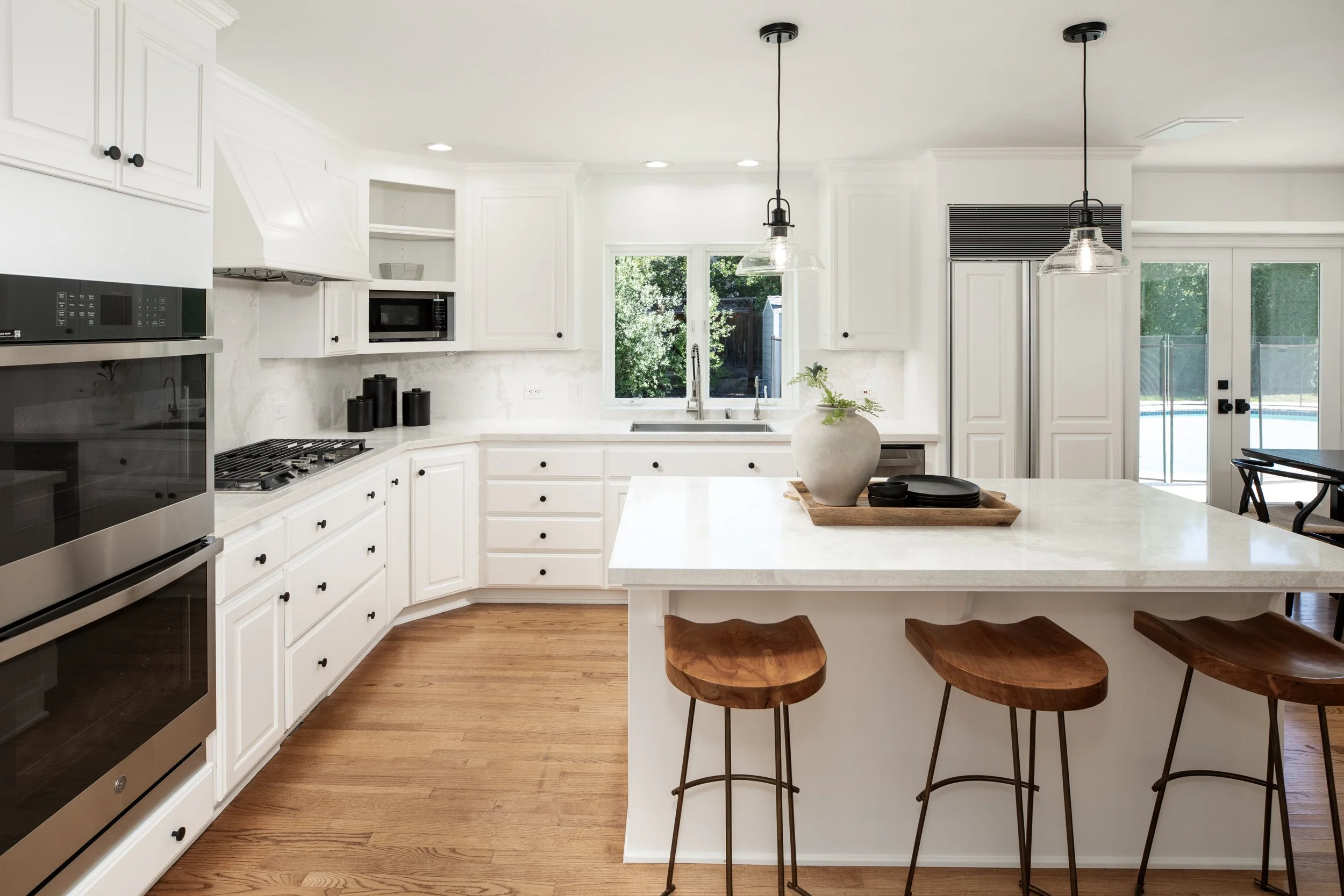 A bright white kitchen with white cabinets, black knobs, and stainless steel appliances, featuring a large island with wooden barstools, pendant lights, and a view of a backyard through a window and a glass door.