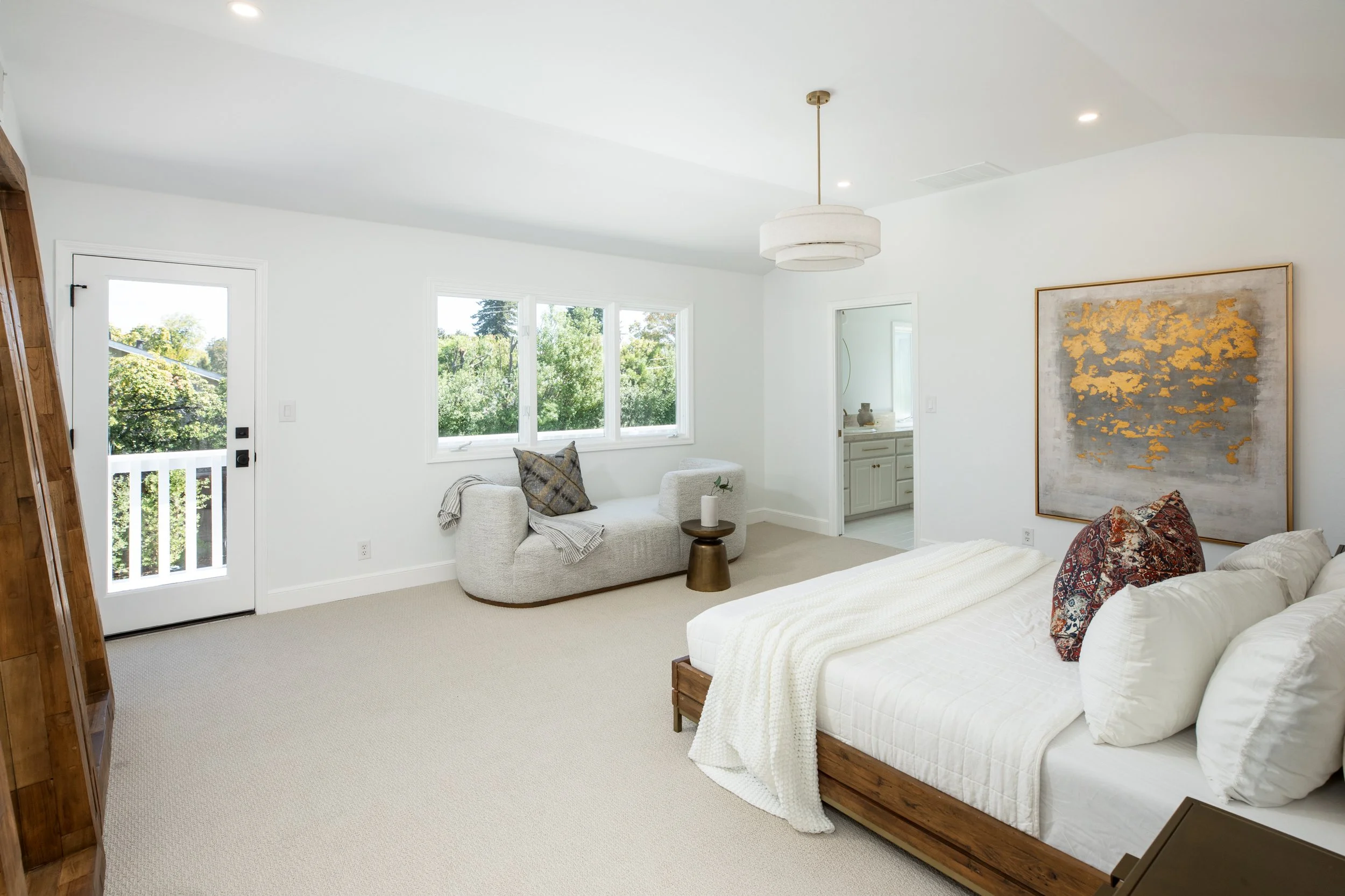 Bright bedroom with a white bed and patterned pillows, a beige armchair by the window, and a large abstract gold and gray painting on the wall. There's a small round side table and windows showing green trees outside.