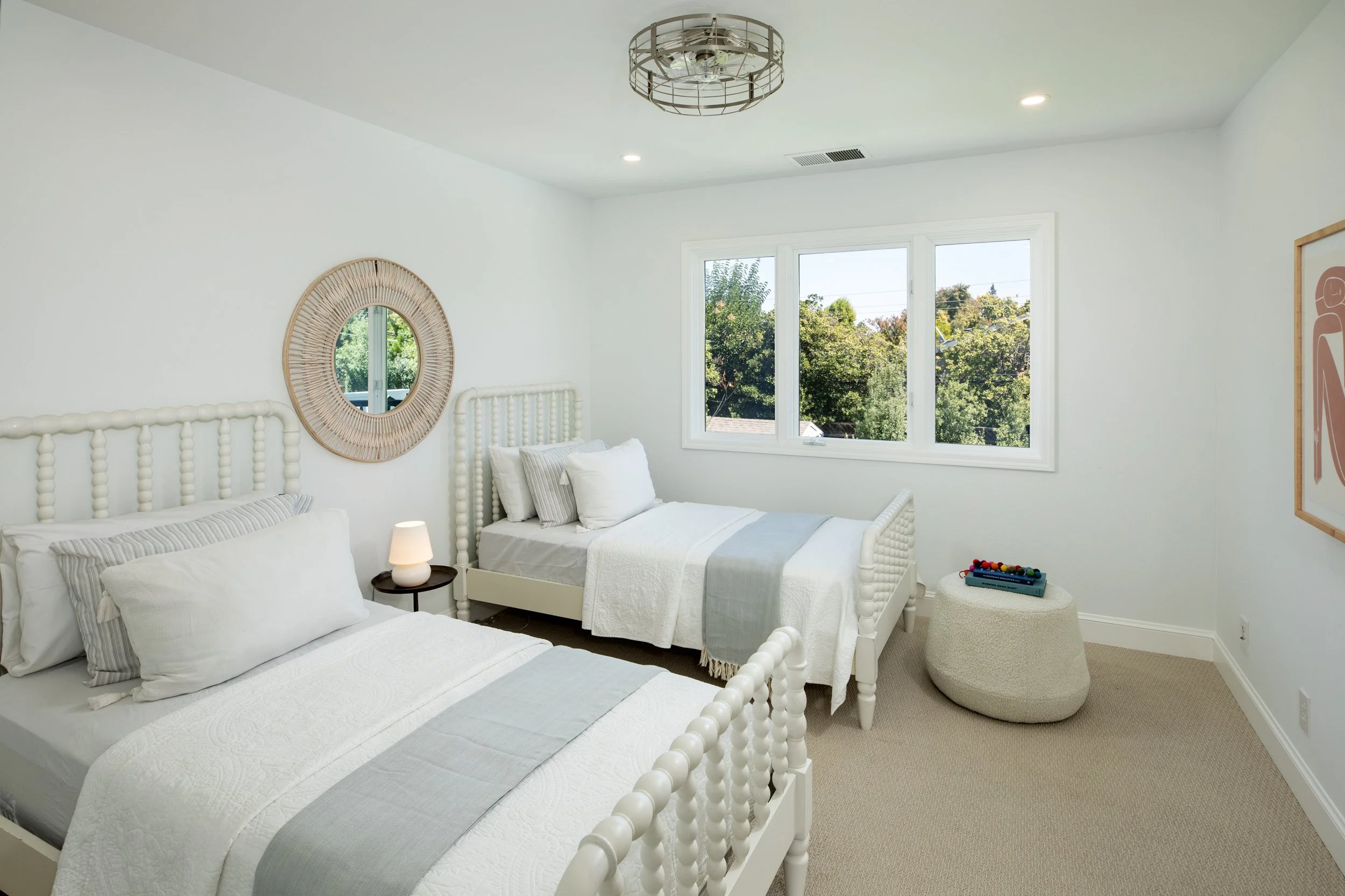 Bright bedroom with two white beds, one with striped pillows, a small round nightstand with a lamp, framed artwork on the wall, a large window showing greenery outside, ceiling light fixture, and neutral-colored carpet.