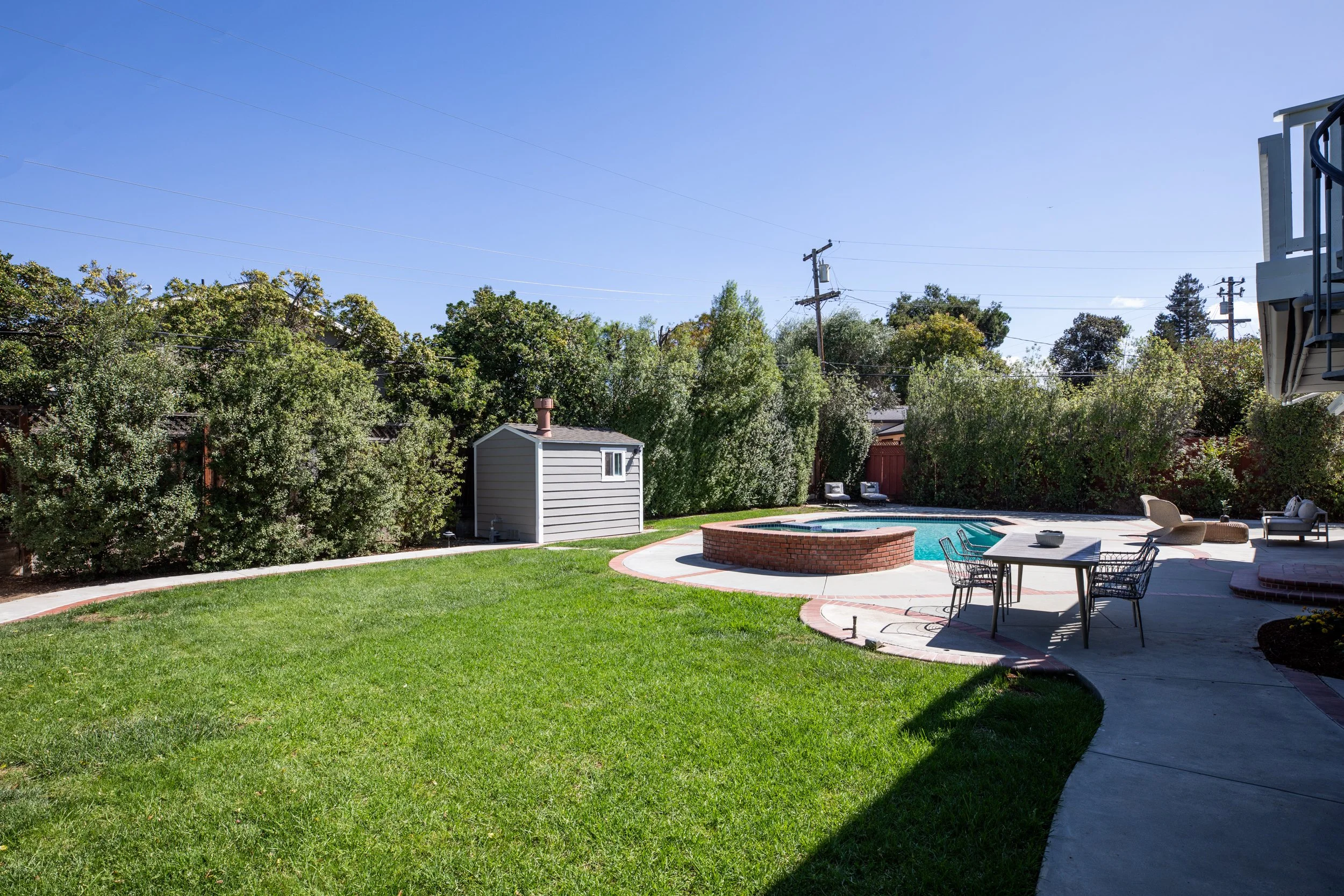 A backyard with a swimming pool, a round brick firepit, and outdoor furniture including chairs and a table. There are trees and bushes along the fence, a small shed, and a clear blue sky overhead.