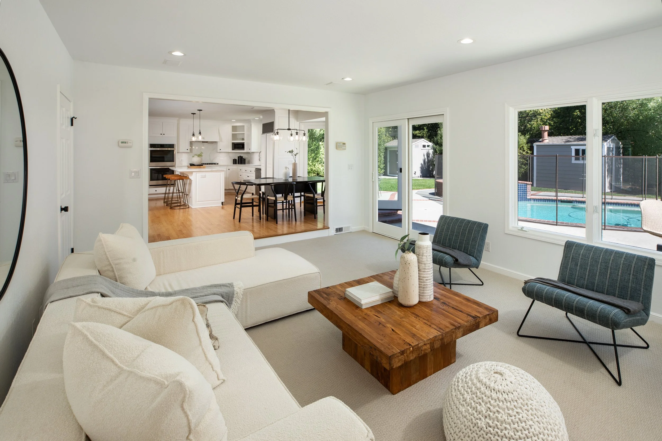 Living room with beige sofa, wooden coffee table, and armchairs, overlooking a kitchen and backyard with a swimming pool through large windows.