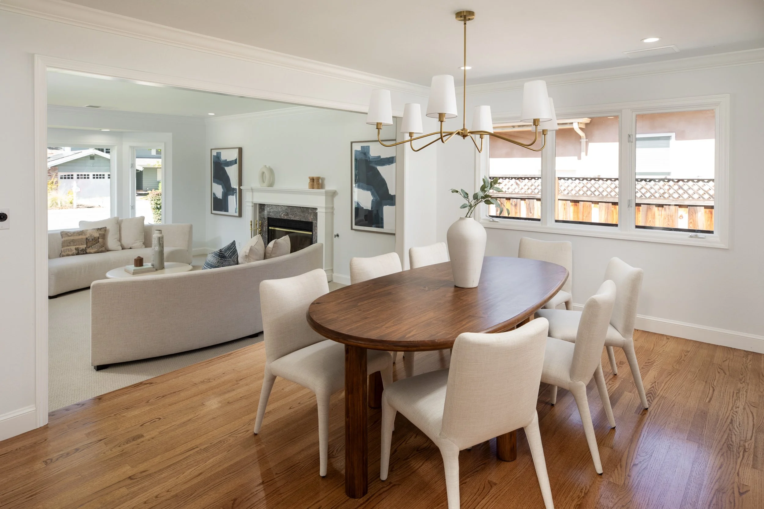 A dining area with a round wooden table and six upholstered chairs, a white vase with greenery, and a window view of a fenced backyard. Adjacent living space includes a cream-colored sofa, artwork, and a fireplace.