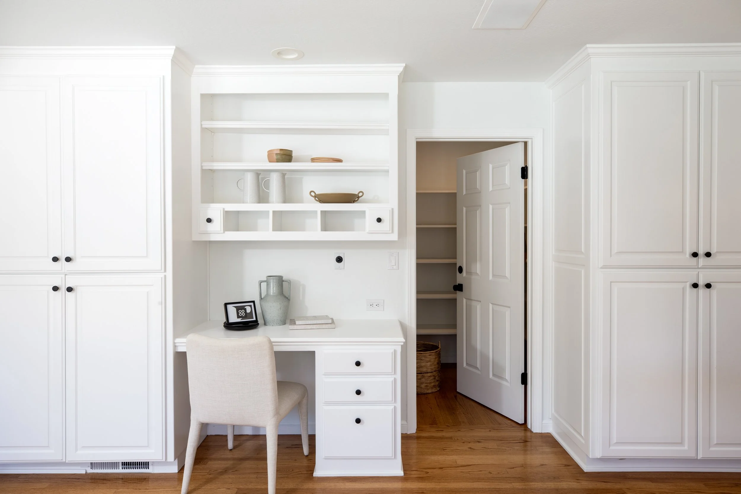 A white built-in desk area with open shelving, drawers, and cabinets, with a beige upholstered chair in front. The desk has a small framed picture, a vase, and some books. To the right, there is a partially open door leading to a closet with shelves.