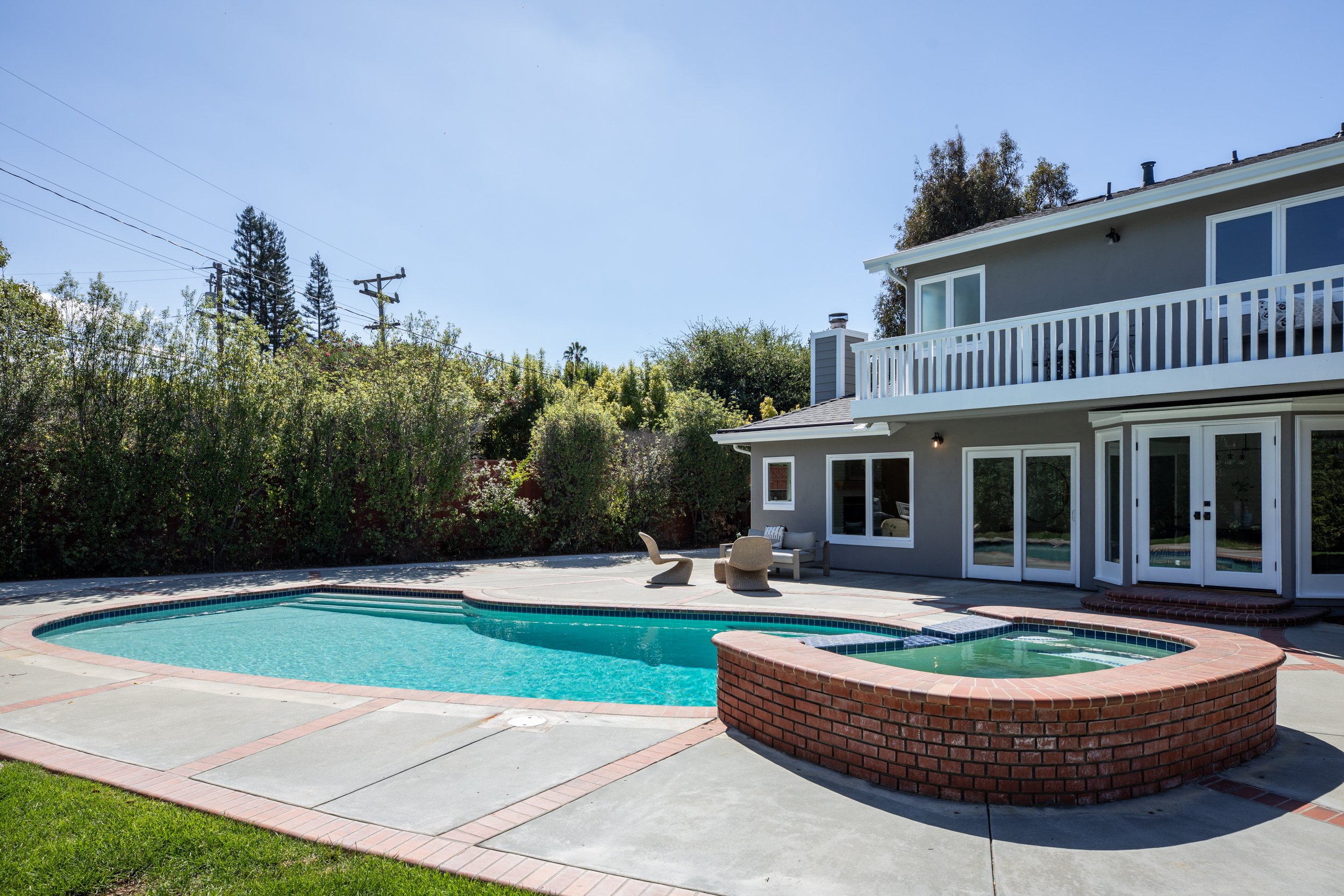 A backyard with a swimming pool, patio furniture, and a two-story house with a balcony, surrounded by greenery and trees under a clear blue sky.