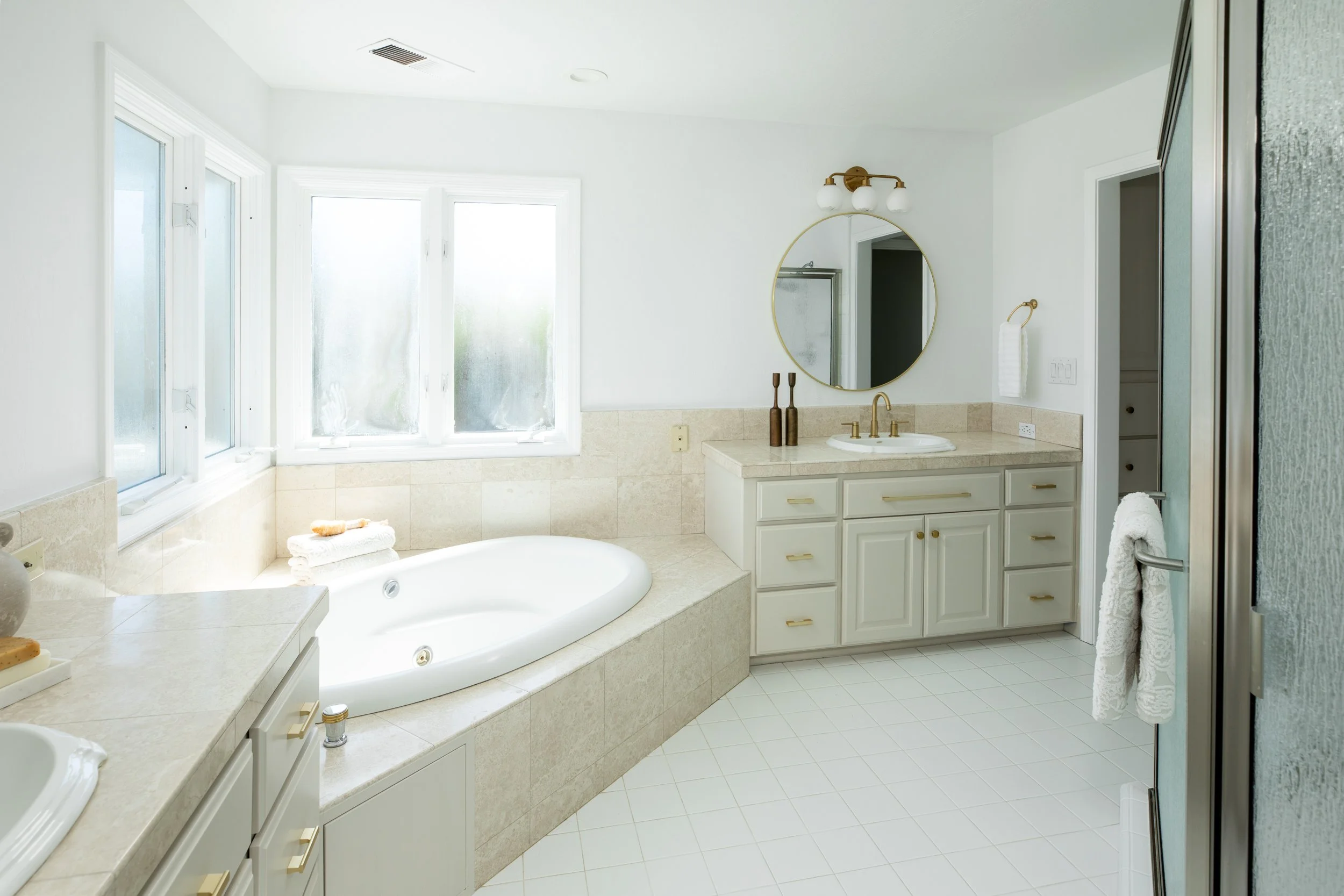 Bright bathroom with large window, white vanity with gold handles, oval mirror, and a whirlpool bathtub with towels on the ledge.