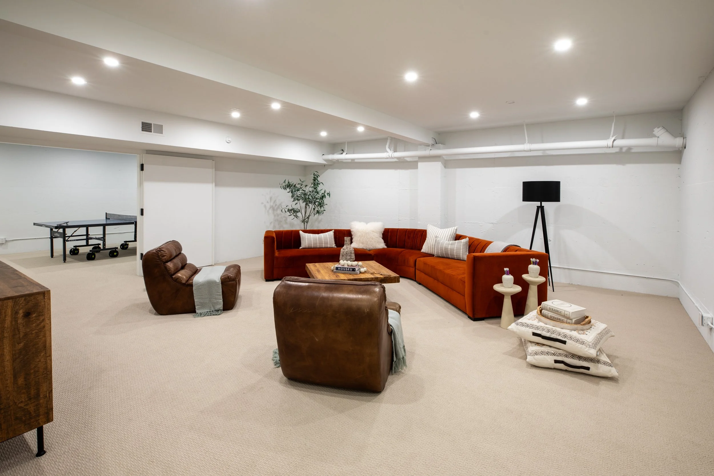 Bright basement living room with white walls and ceiling, beige carpet, orange velvet sectional sofa with decorative pillows, two brown leather armchairs, coffee table with decorative items, and a ping pong table in the corner.