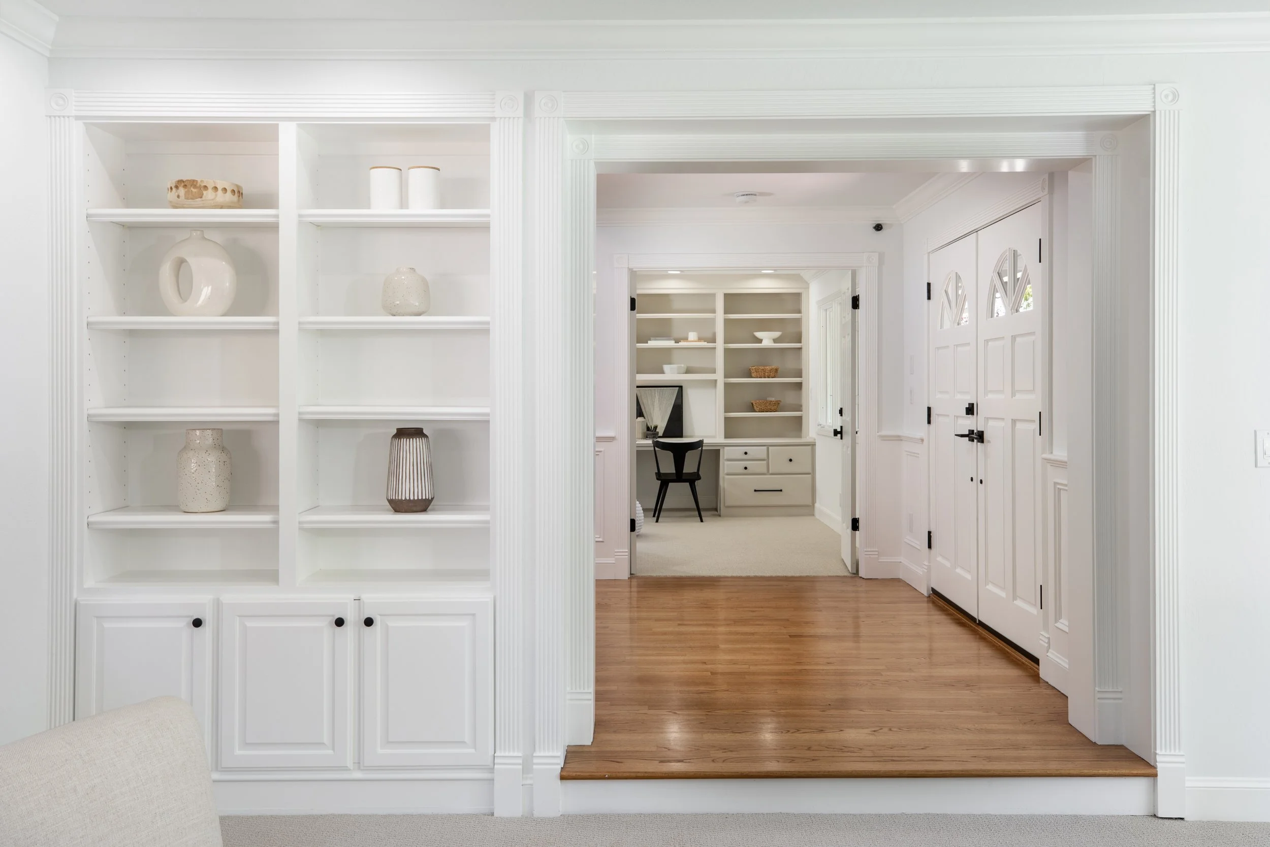 Interior view of a white built-in bookshelf with decorative vases on the shelves, leading to a room with more shelves and a desk, and a wooden floor.