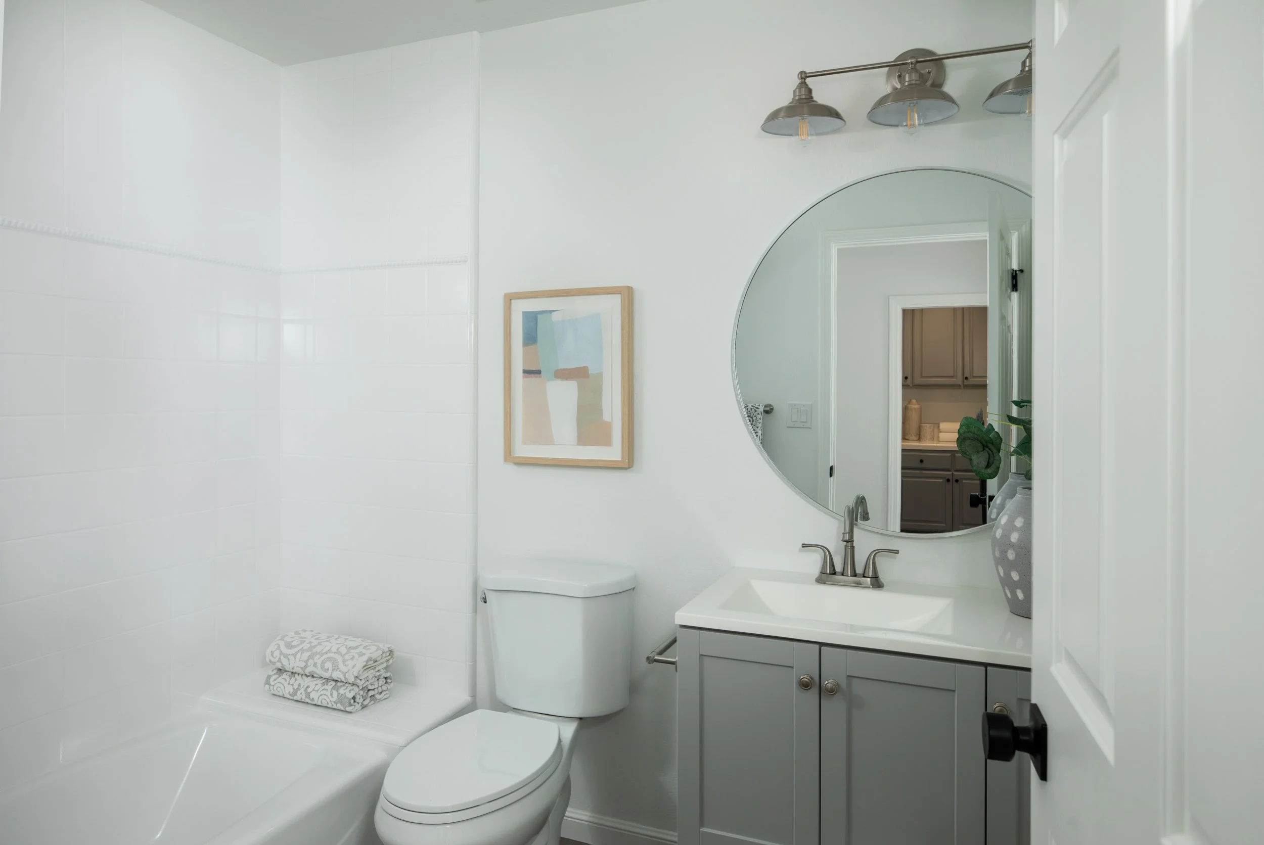 A modern bathroom featuring a white toilet, a round mirror above the sink, a small cabinet, a framed abstract artwork, and a stack of folded towels on the bathtub ledge. The wall has a metallic light fixture with three shades.