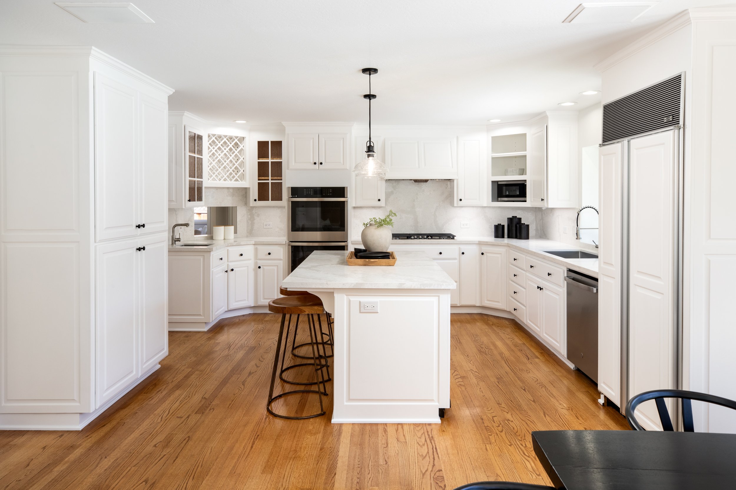 Bright white kitchen with wooden flooring, white cabinets, a kitchen island with a potted plant, built-in oven, microwave, and refrigerator, pendant light over island, black bar stools, and black dining table with chairs.