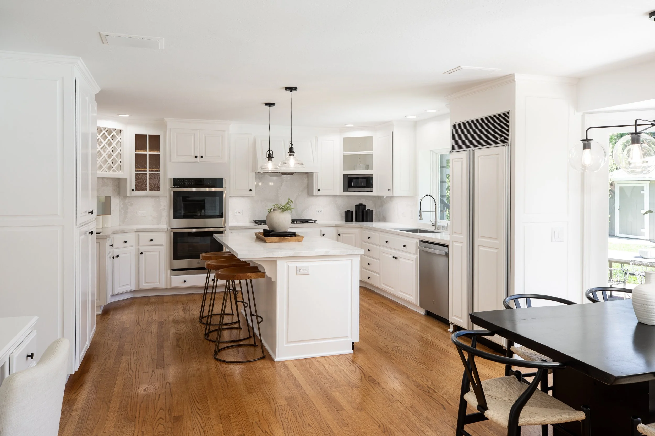 Modern white kitchen with wooden flooring, island with wooden stools, and dining table with black chairs, natural light from windows, pendant lights, built-in appliances, and decorative vases.