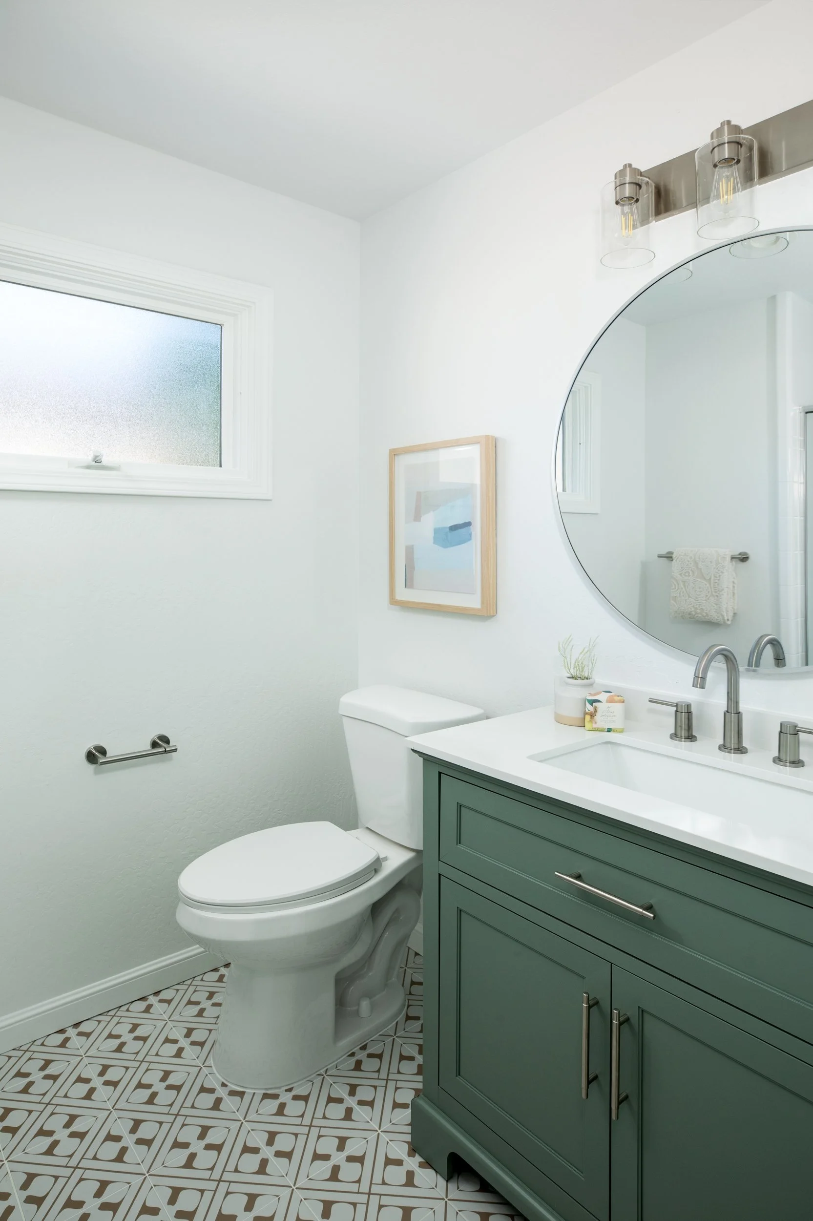 A modern bathroom with white walls, a small window, a white toilet, a green vanity with a white countertop and silver hardware, a large round mirror, a framed artwork, a potted plant, a soap dispenser, a hand soap container, a towel on a silver towel