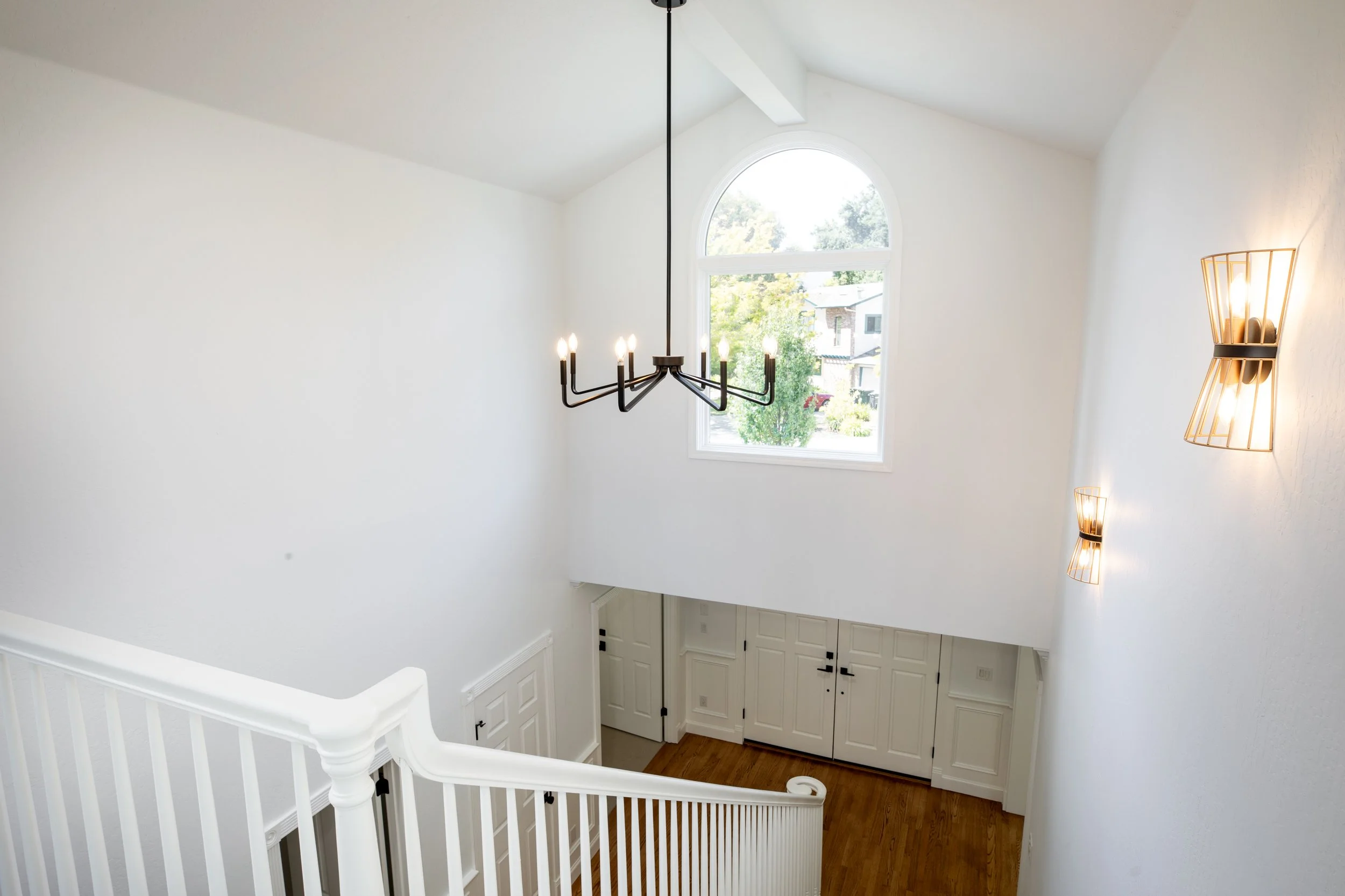 Interior view of a staircase landing with a large arched window, modern chandelier, wall sconces, white walls, and wooden flooring.
