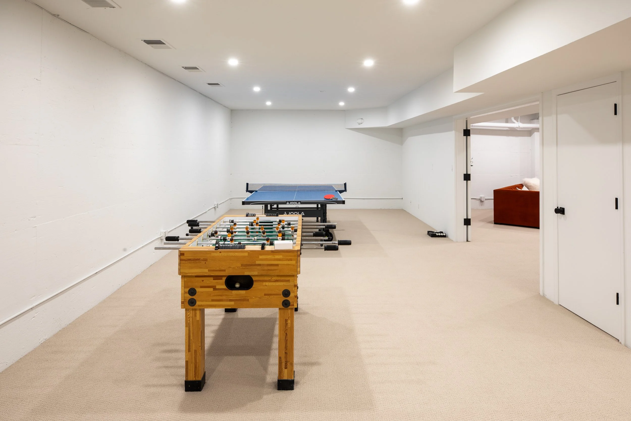 A game room with a foosball table and a ping pong table in a spacious, well-lit room with white walls and beige carpet.