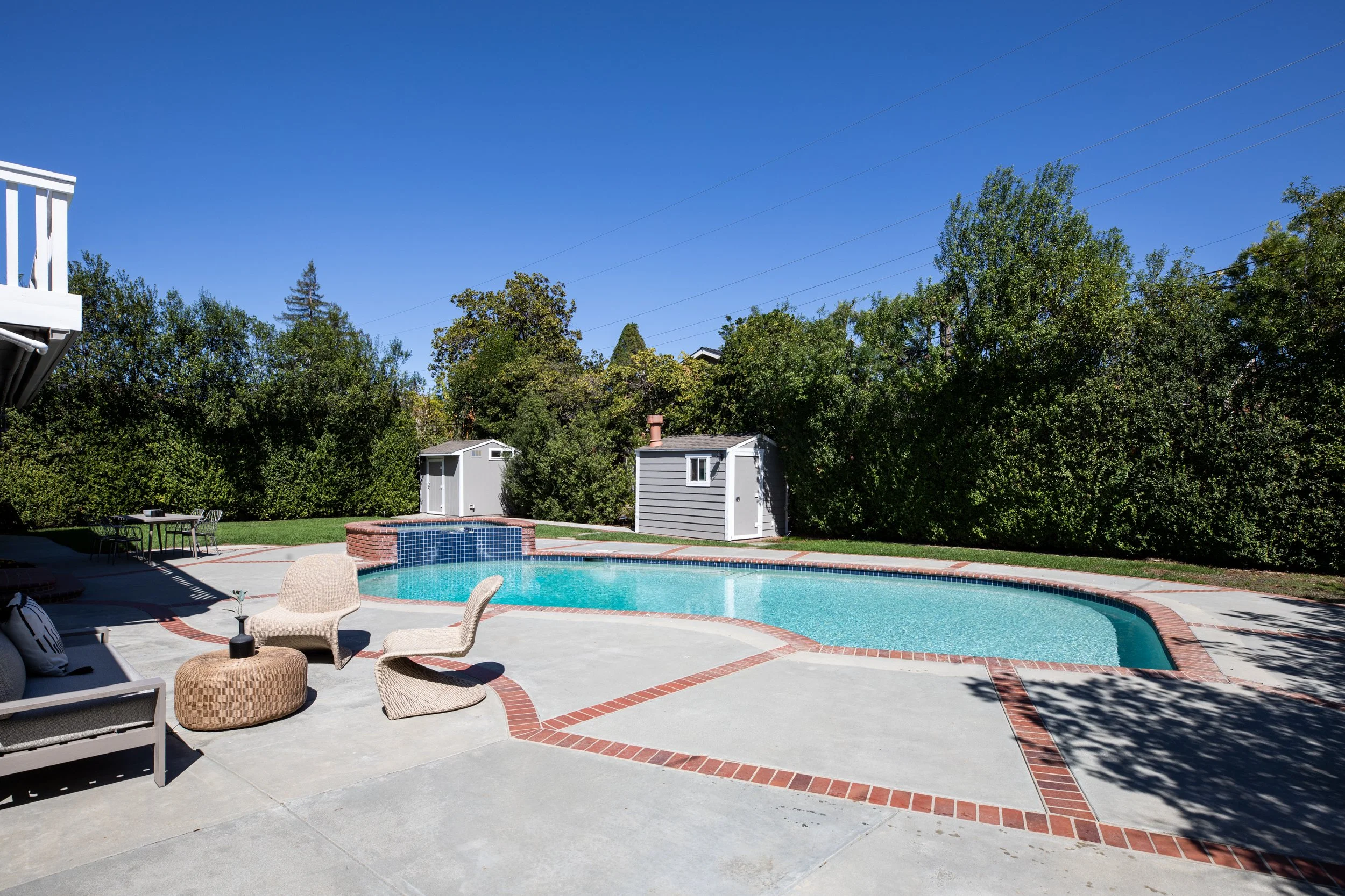 A backyard with a swimming pool, outdoor furniture, a small shed, trees, and a clear blue sky.