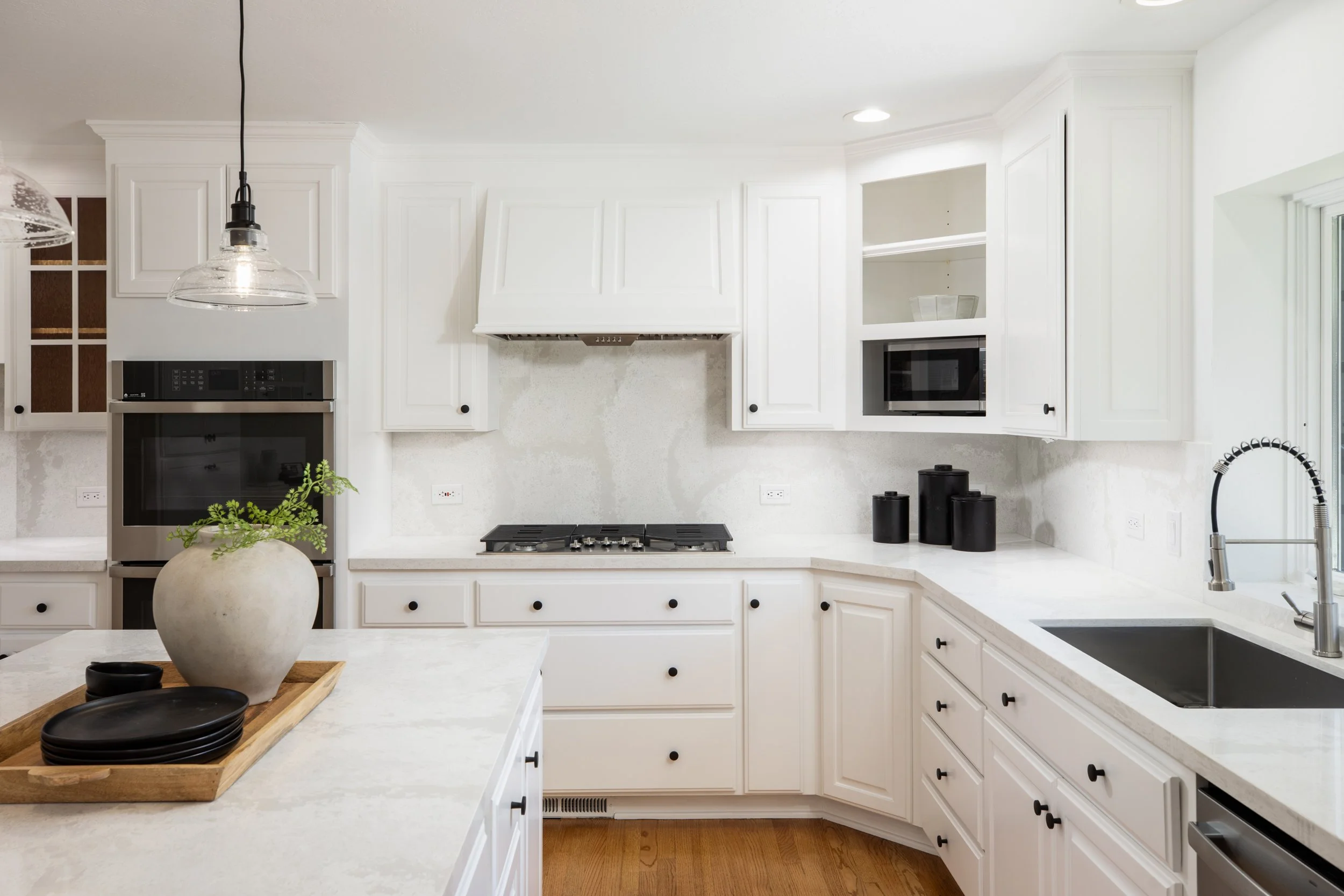 A white kitchen with cabinets, a microwave, a built-in oven, a stovetop, and a modern pull-down faucet. There are black knobs on the cabinets, a white countertop, and a potted plant on the island. Recessed lighting and a hanging pendant light are als