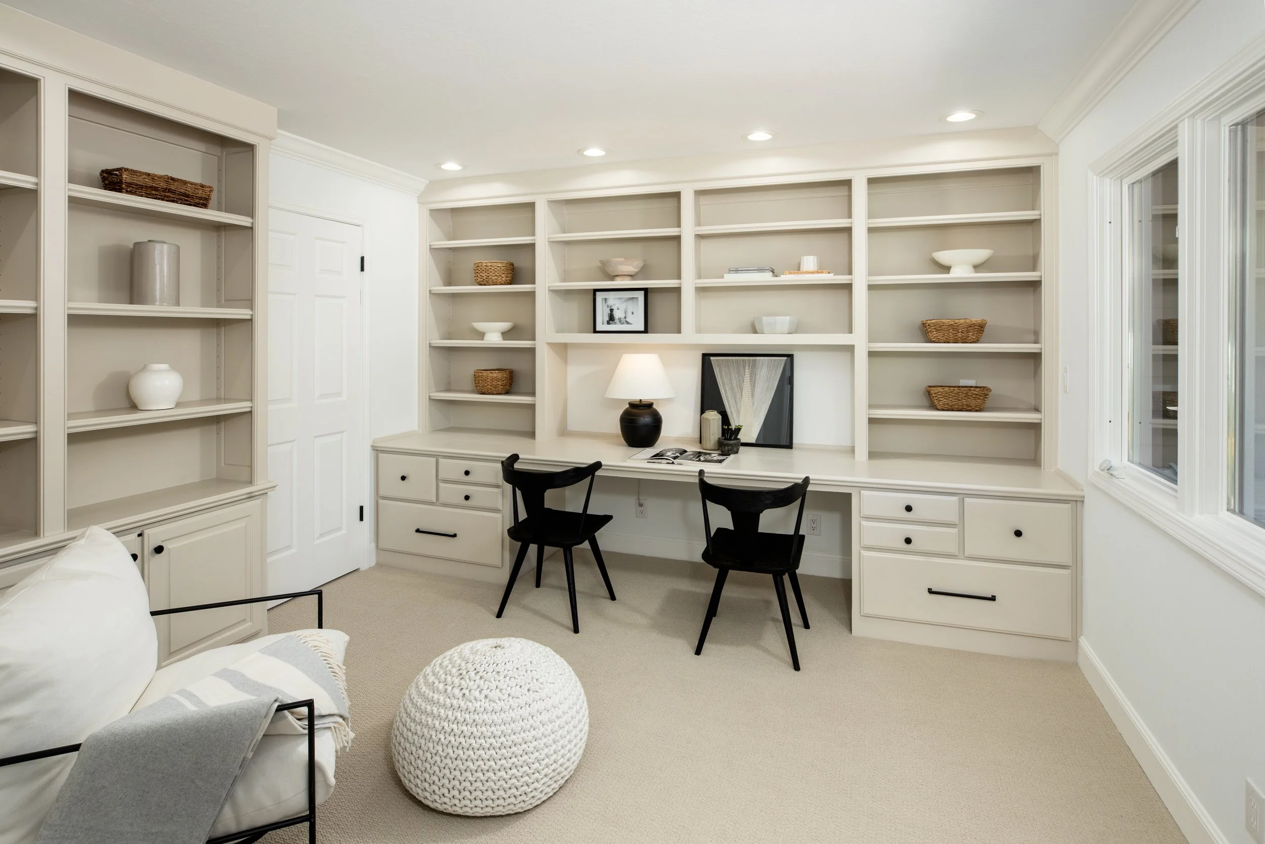 A cozy home office with built-in white shelving, a desk with a table lamp, two black chairs, a framed photo, and decorative bowls and baskets, with a window on the right and a bed partially visible in the foreground.