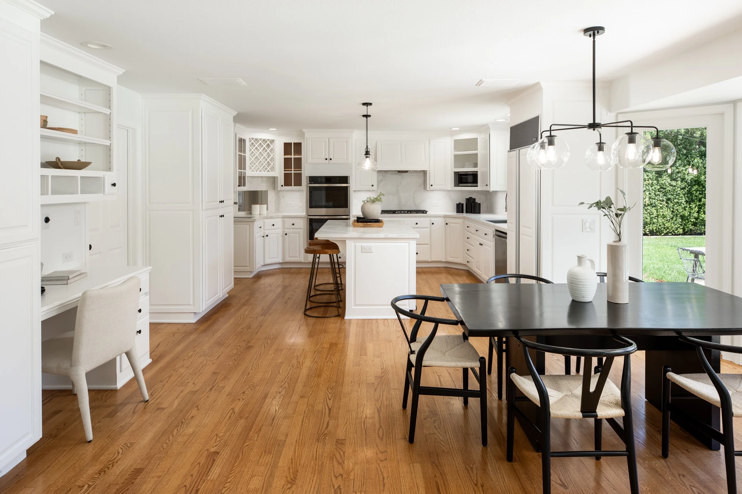 Bright, modern kitchen with white cabinetry, marble countertops, wooden flooring, and black fixtures. There is a kitchen island with two stools, a dining table with black chairs, and decorative vases and plants. Large windows provide natural light an