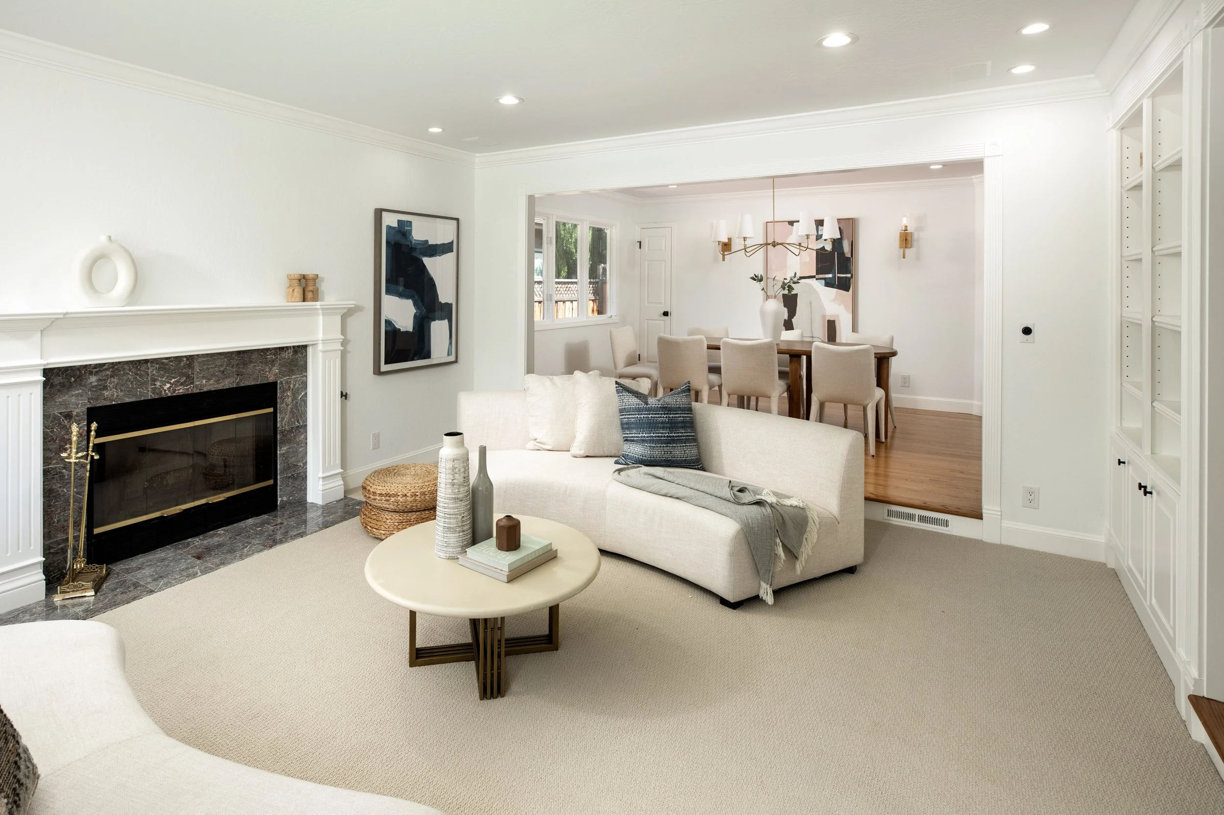 Living room with fireplace, abstract artwork, beige sofa with pillows, round coffee table with vases, staircase, and dining area in the background with a chandelier and additional artwork.
