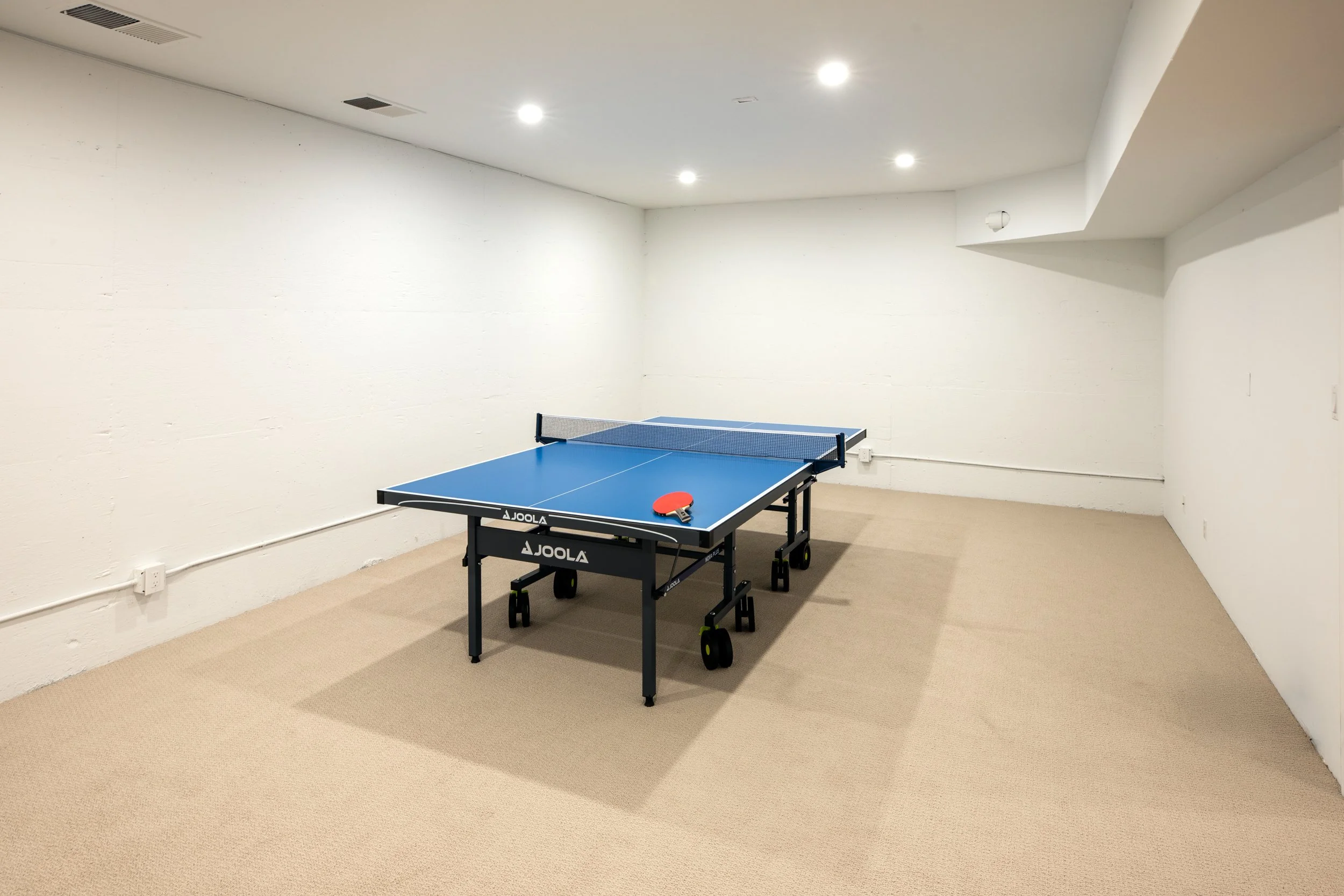 An indoor room with beige carpeted floor, white walls, and four ceiling lights. There is a blue Joola table tennis table with a red paddle and a net in the center of the room.