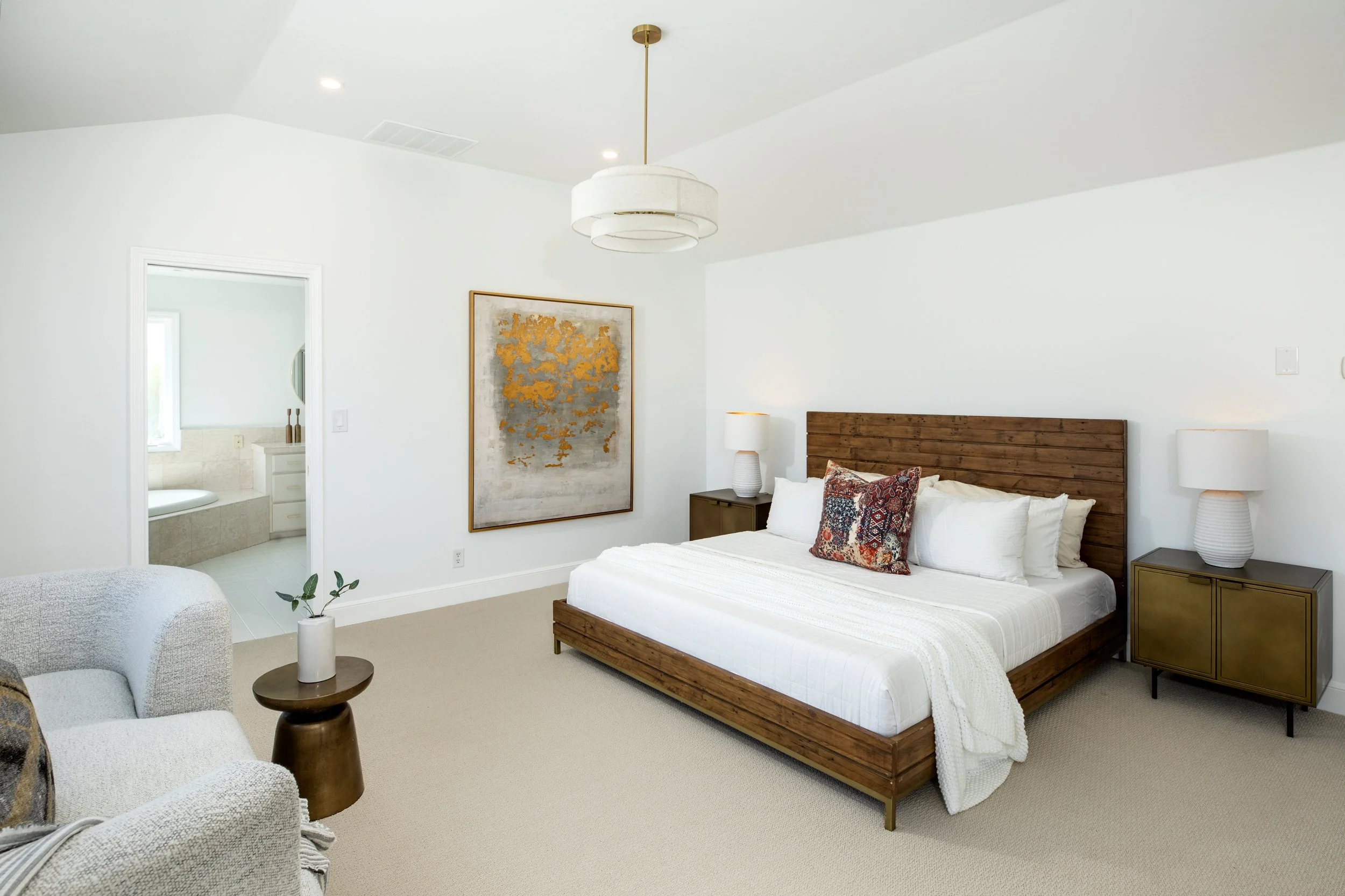 A bedroom featuring a bed with a wooden headboard and white bedding, two bedside tables with lamps, abstract artwork on the wall, a seated area with a white armchair, a small table with a plant, and an attached bathroom visible through an open door.