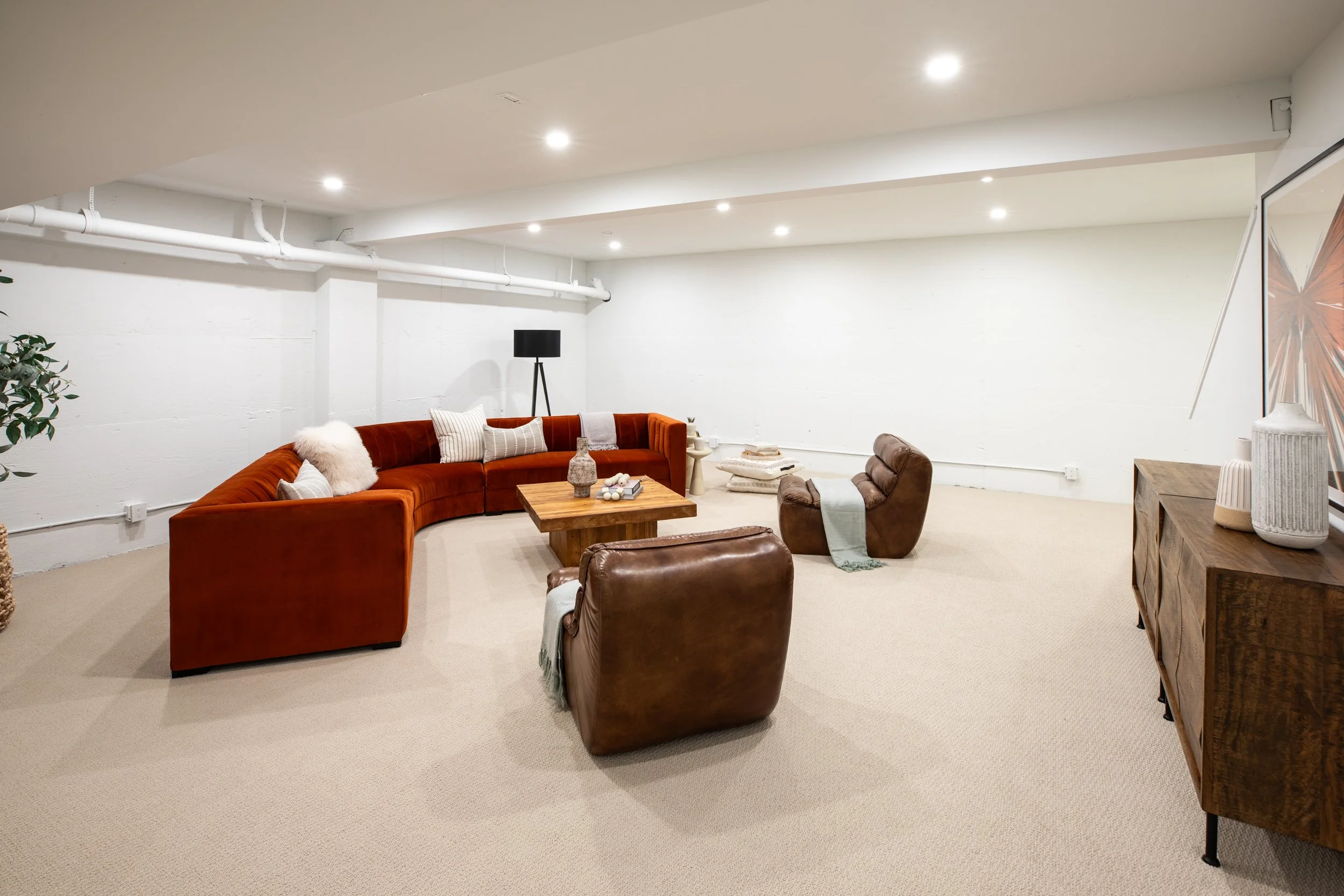 A spacious basement living room with white walls and beige carpet. Features a curved rust-colored velvet sofa with throw pillows, two brown leather armchairs, a wooden coffee table with decorative items, a black floor lamp, and a large framed butterf